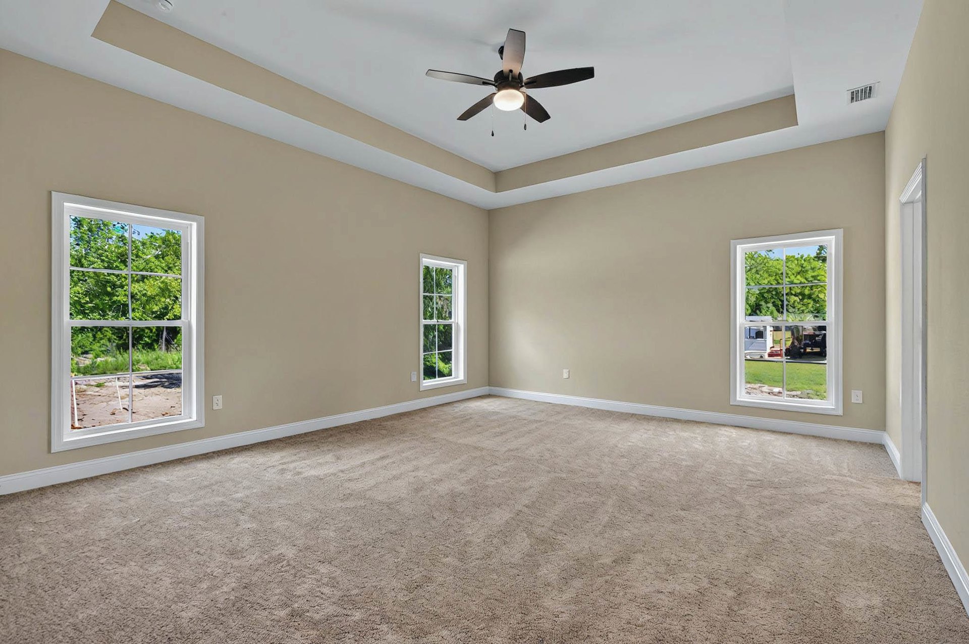 Carpeted room with white-framed windows overlooking trees and lawn, ceiling fan with light fixture, neutral walls and ceiling