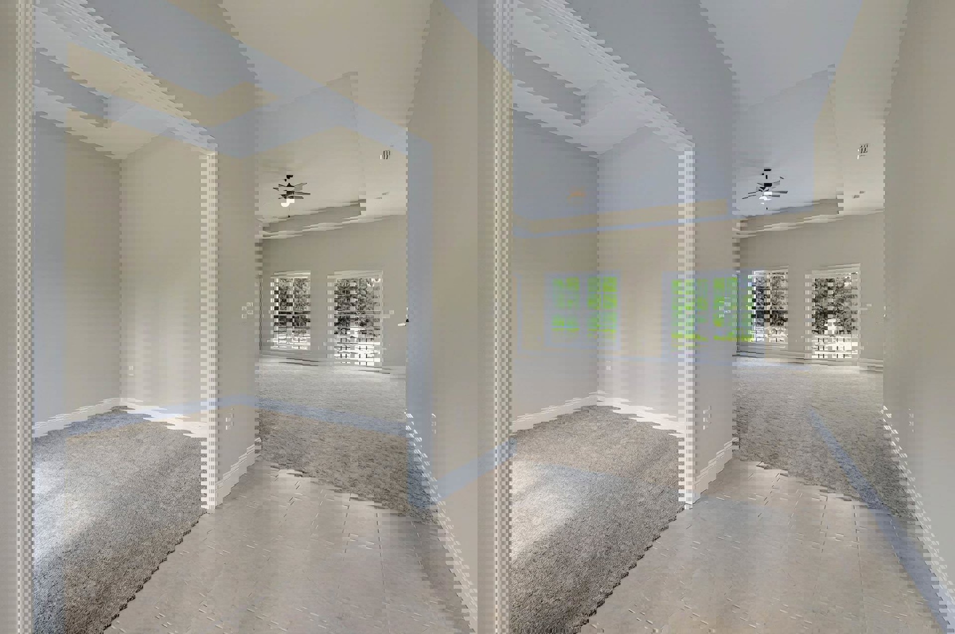 Beige walls and carpeted floor, ceiling fan with light fixture, large window overlooking trees, white tile floor partially covered by a rug