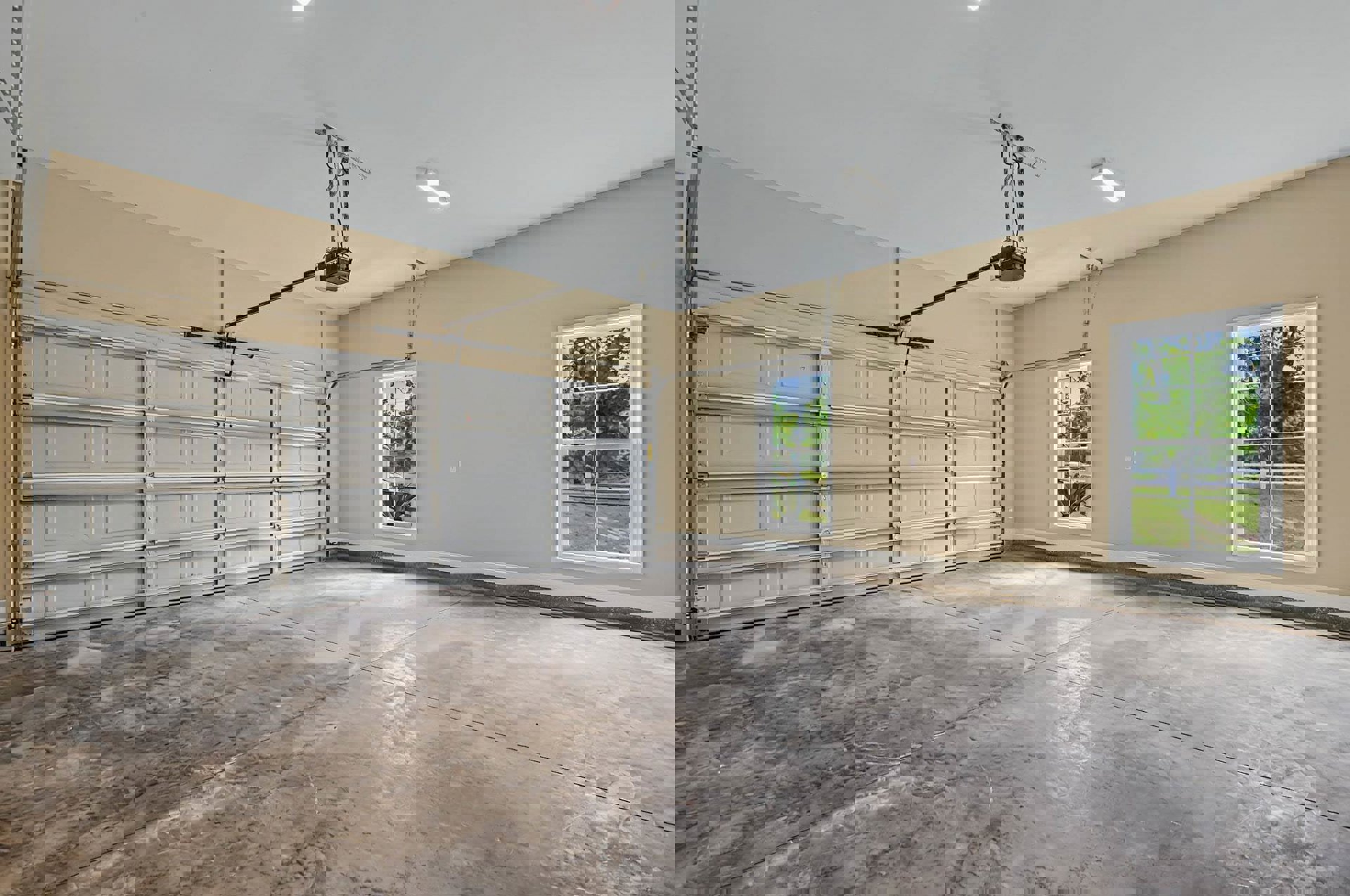 Spacious garage with concrete floor, white walls, ceiling light fixture, metal-framed garage door with pulley, and windows overlooking lawn and trees