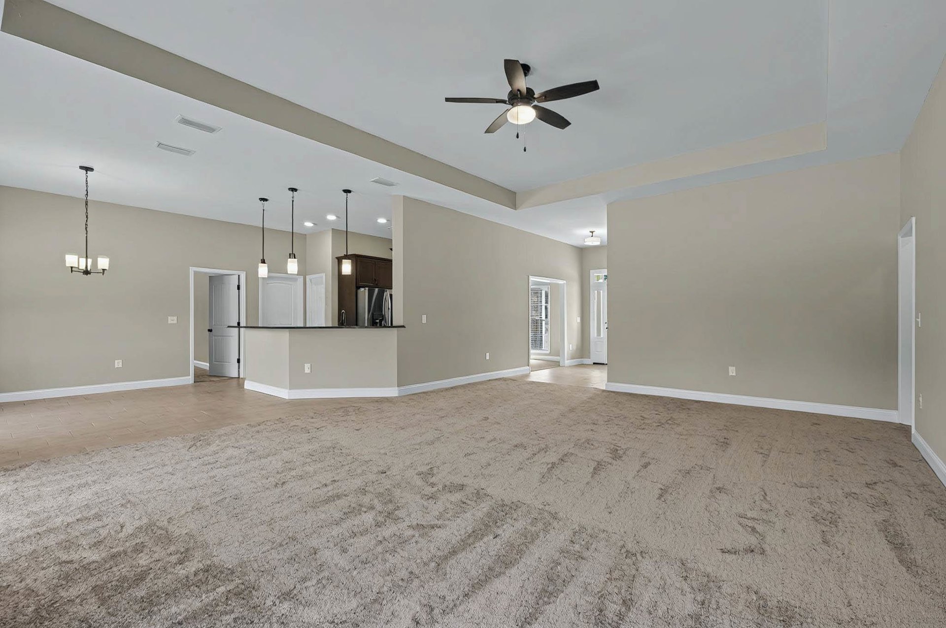 Open living room with carpeted floor, ceiling fan with light, white door with black knobs, window with white shutters, and adjacent kitchen area