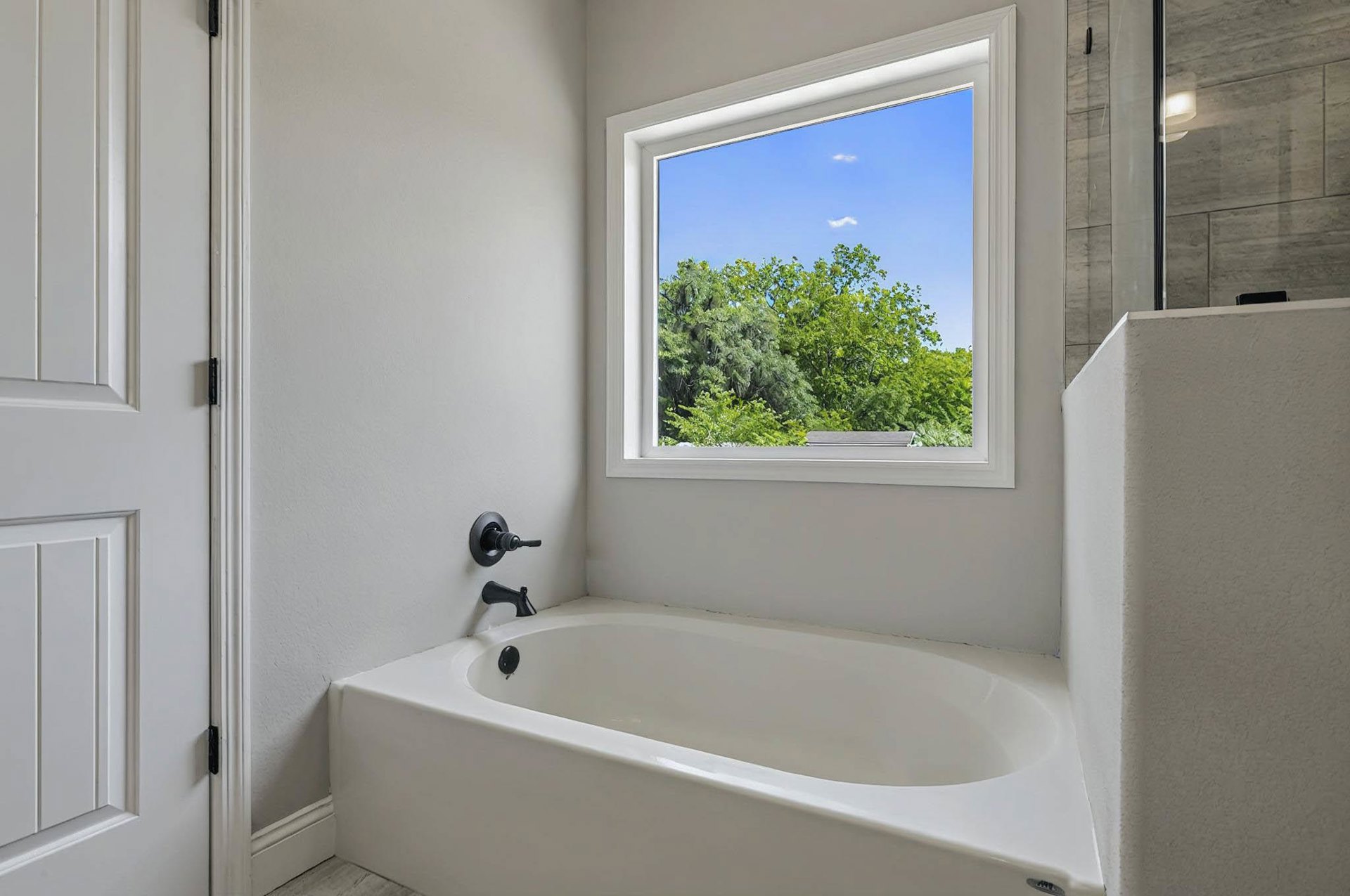 Freestanding white bathtub beneath large window with view of green trees and blue sky, chrome faucet, white walls, bathroom interior