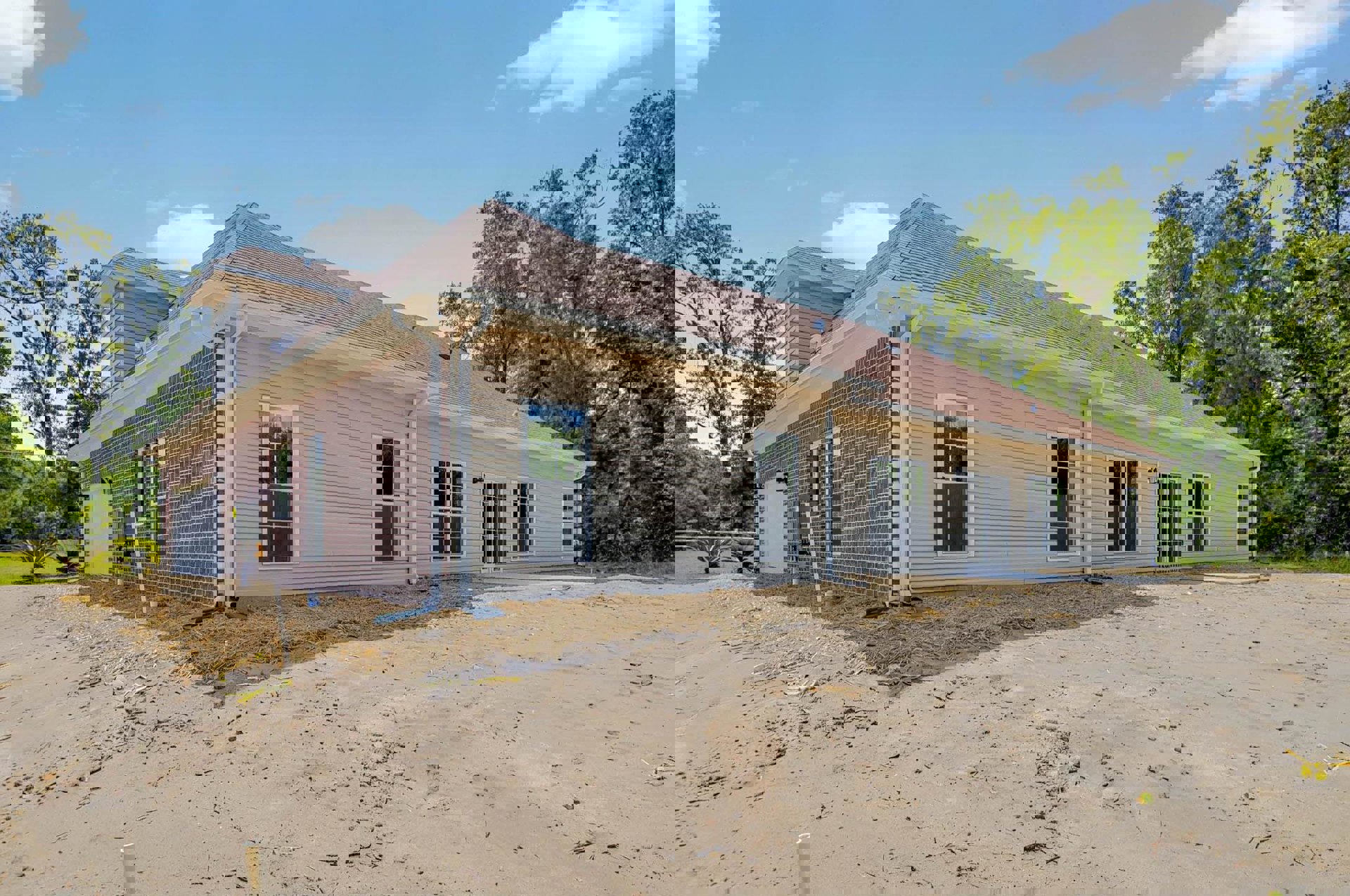 Partially built house with white-framed windows, exposed roof, dirt-covered ground, and surrounding trees under a cloudy sky