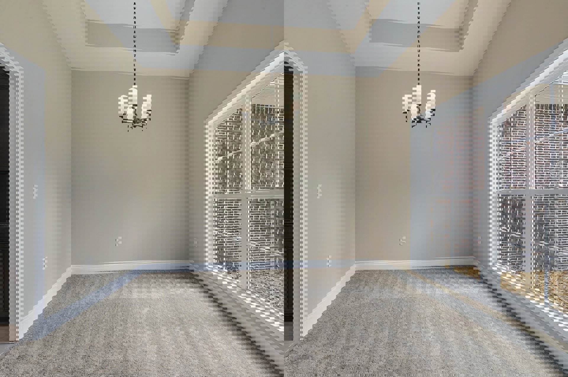 Spacious room with white walls, black door, large window set in a brick accent wall, plush carpet flooring, and elegant chandelier with white lampshades hanging from molded ceiling