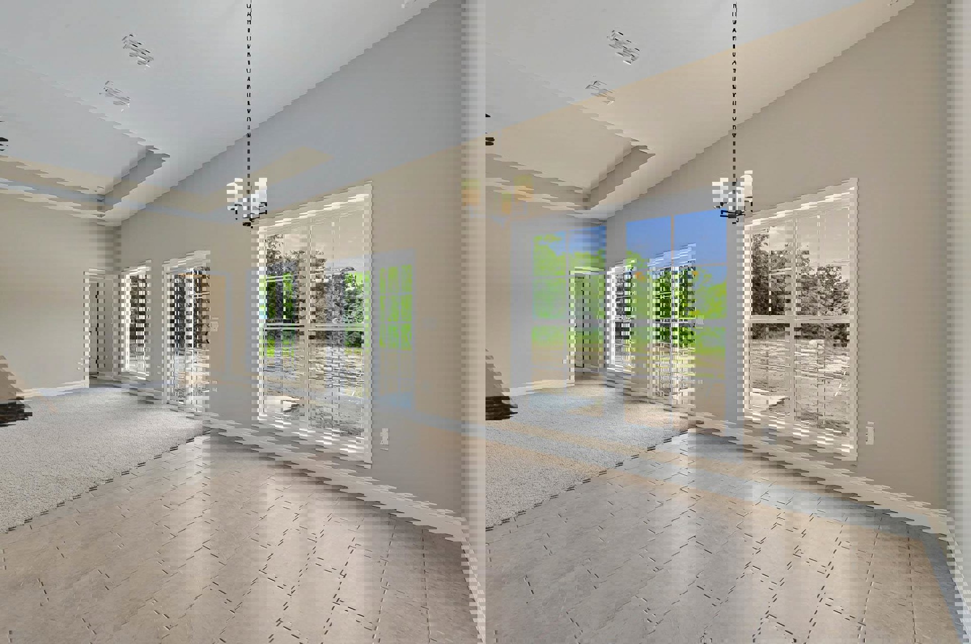 Carpeted room with large windows, white window frames, and view of trees outside; neutral walls and ceiling, tile flooring visible near entry.