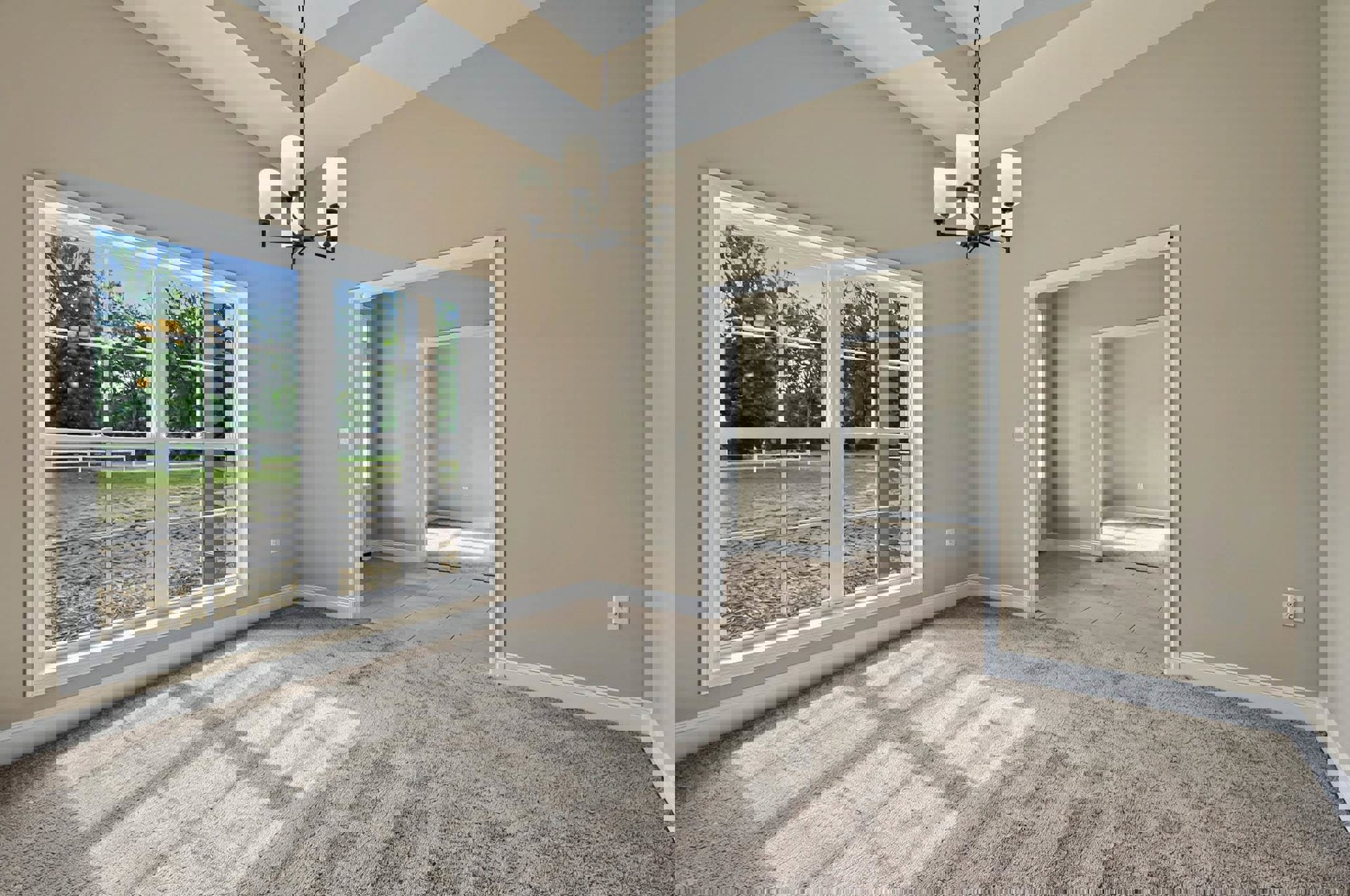 Spacious room with large windows overlooking a field and trees, ornate chandelier hanging from a plaster ceiling, neutral carpet flooring, white walls with decorative molding