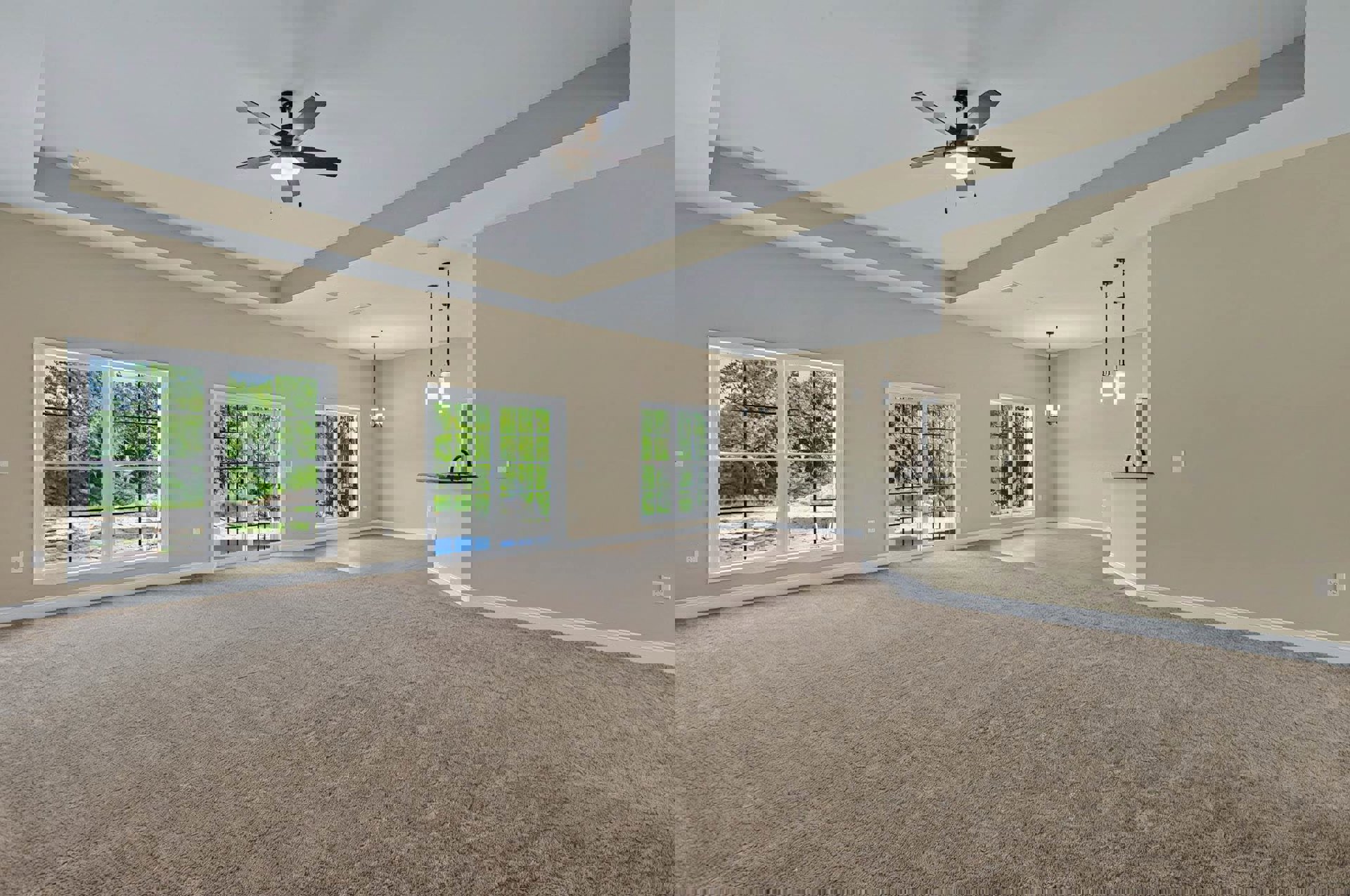 Carpeted living room with ceiling fan and light, multiple windows showing leafy trees, white walls, and doorway leading to kitchen