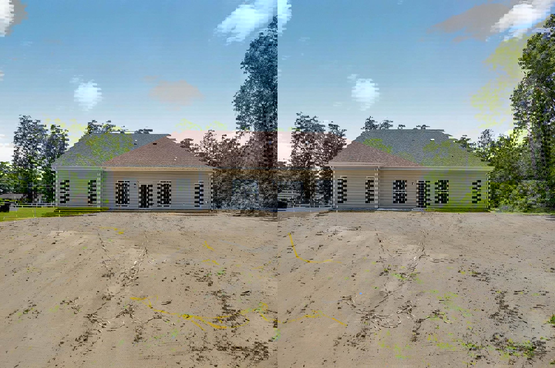 Modern house with gray roof and white-framed windows, surrounded by dirt and sand, yellow caution tape on ground, trees and cloudy sky in background