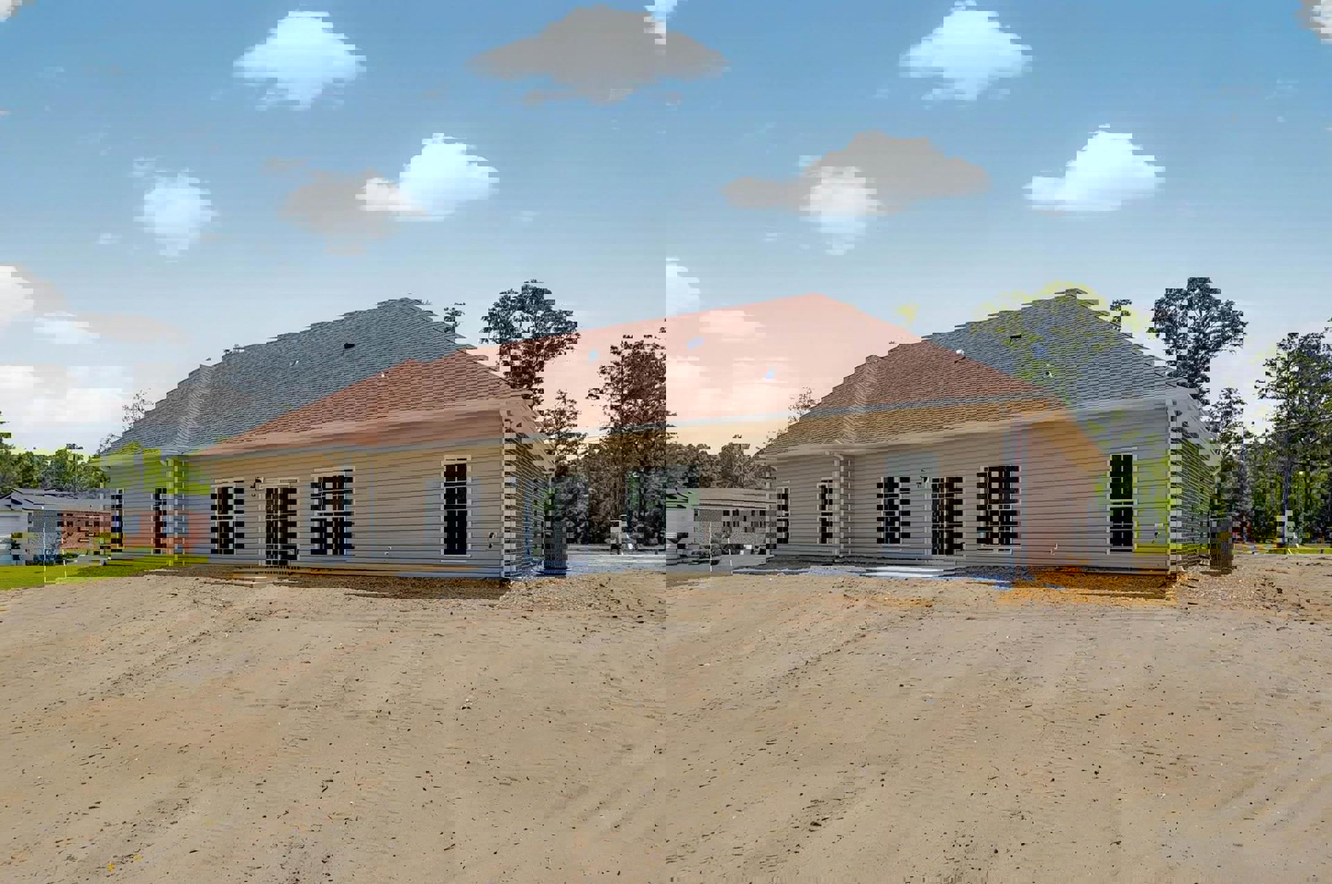 Partially built house with exposed framing, shingled roof, white-framed windows, dirt driveway, surrounding trees, and cloudy sky