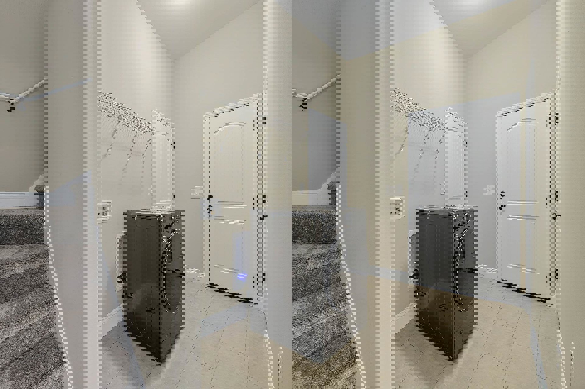 Laundry room with black washing machine on light tile floor, white door with black handle, white metal shelf with hanging rods, white walls and ceiling