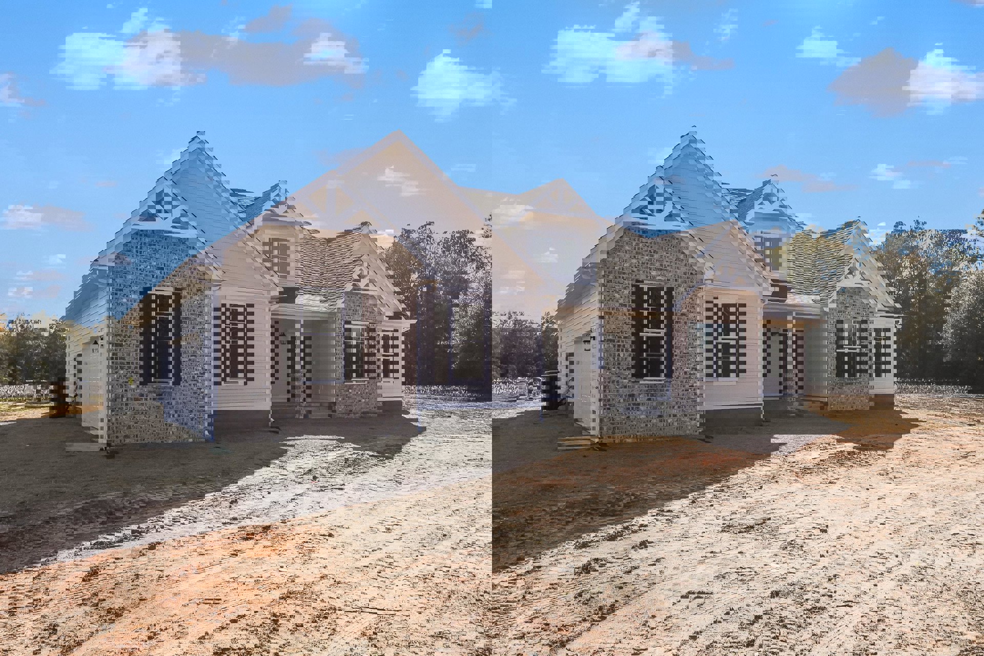 Two-story house with white-framed windows, gray roof, and dirt driveway, surrounded by trees and cloudy sky; black rectangular object with yellow label near the entrance.