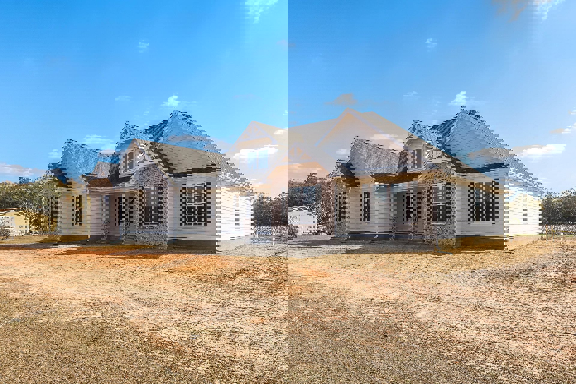 Partially built house with brick and stone exterior walls, dark roof, large window, dirt road and bush in foreground, blue sky with scattered clouds