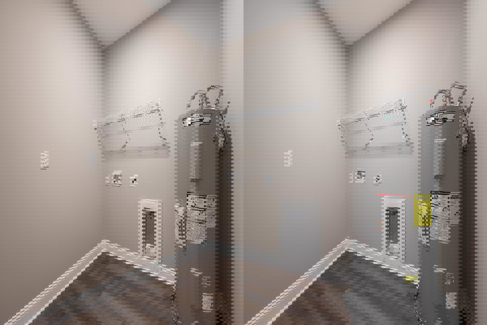 Utility room with a large grey water heater featuring a yellow warning sign, white electrical outlet on plaster wall, silver urinal covered with a plastic bag, white wall shelf