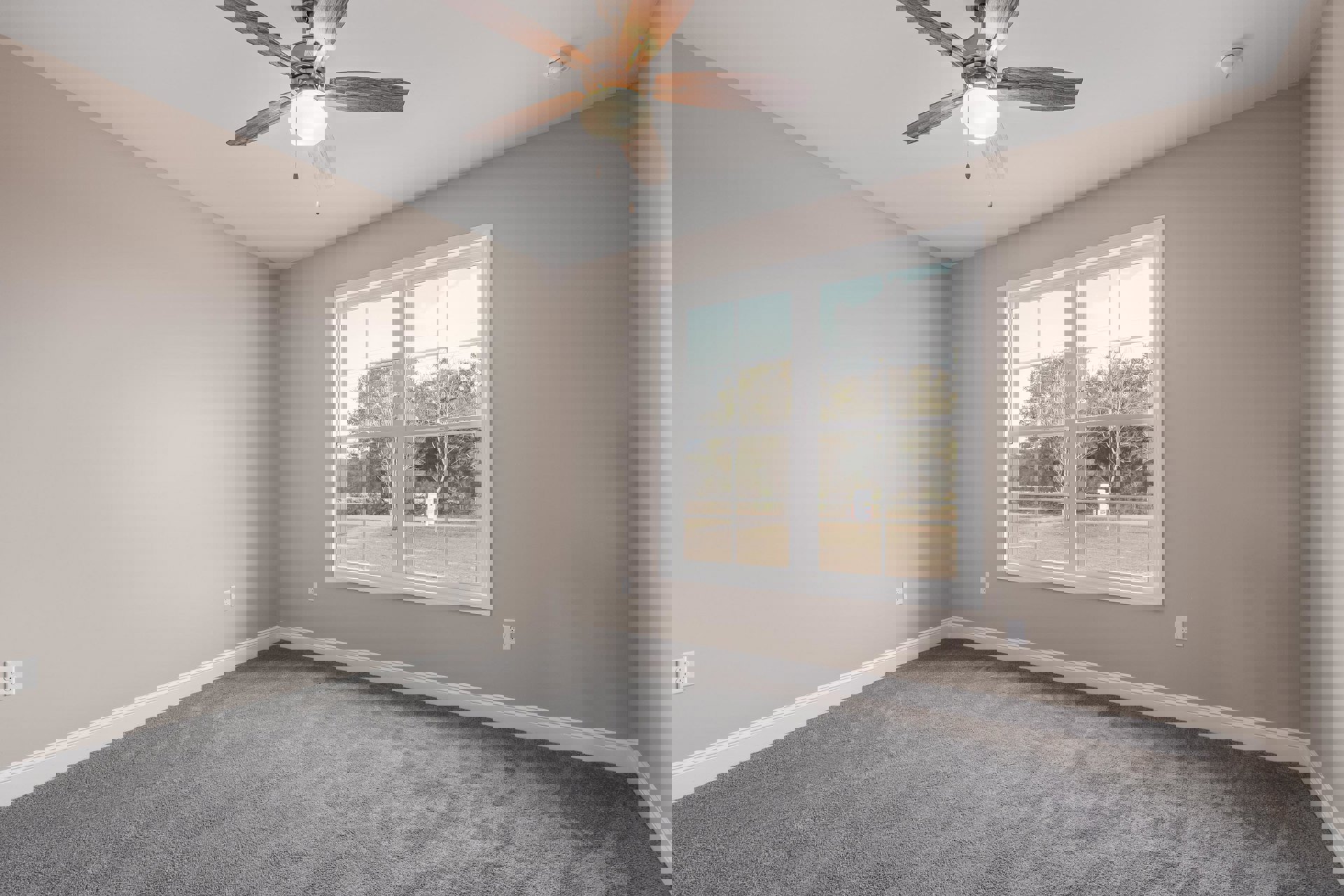 Ceiling fan with light fixture mounted on white ceiling, smoke detector nearby, large window with view of green trees, grey carpet flooring, pale walls with simple molding