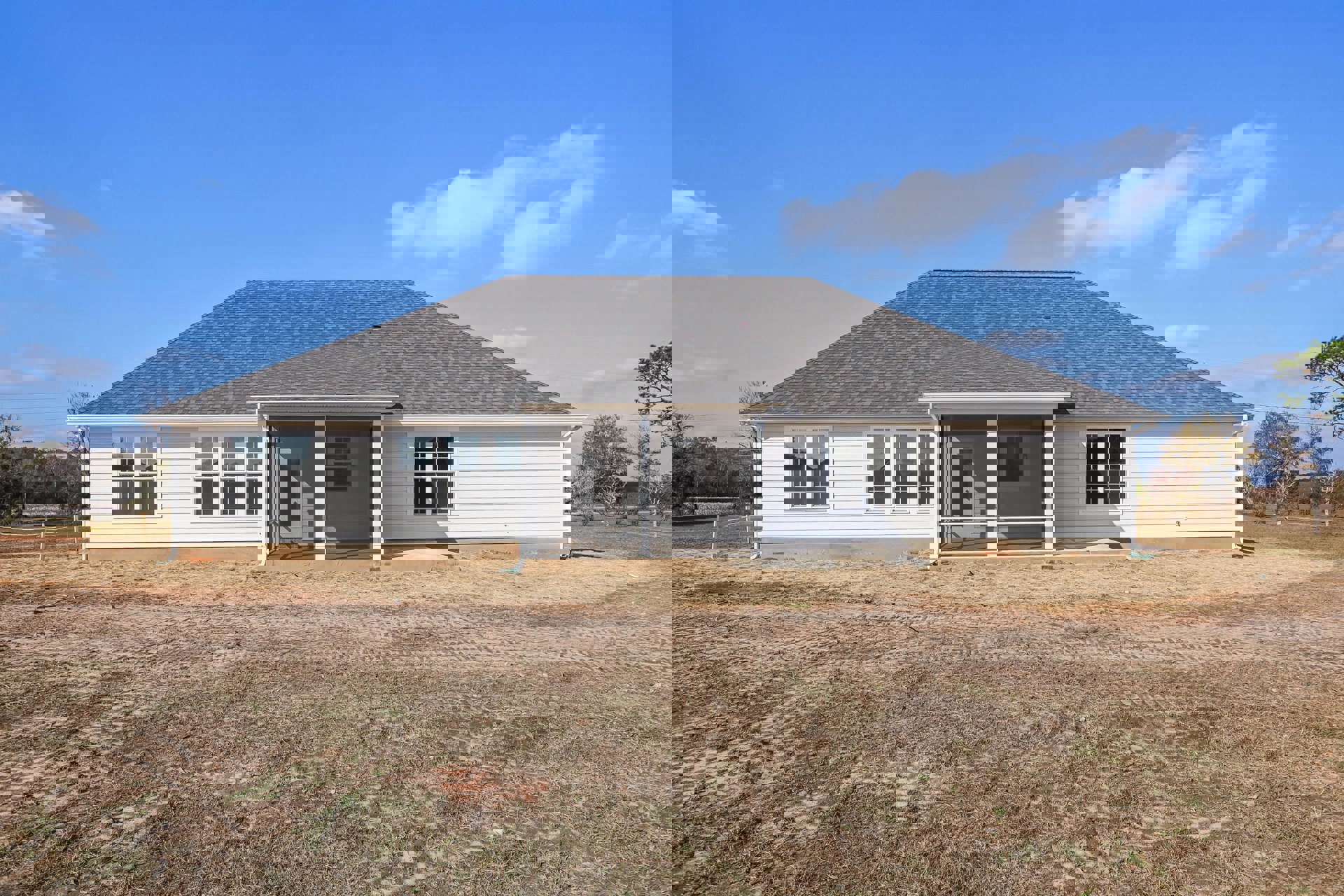 White cottage-style home with screened front porch, multiple square-paned windows, gray roof, expansive grassy yard, dirt driveway with tire tracks, surrounded by trees under a