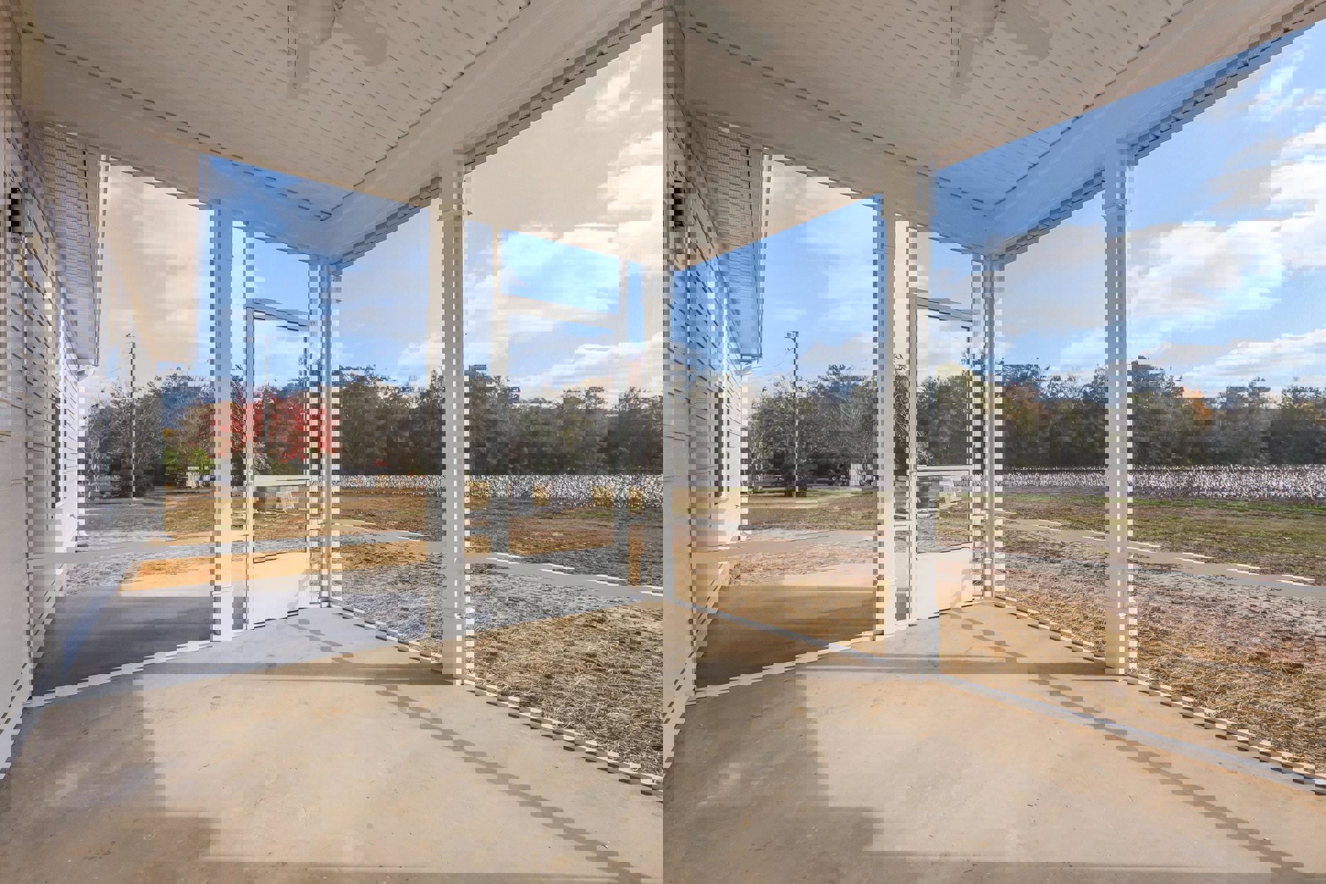 White porch with glass doors, concrete floor casting shadow, overlooking grassy field and trees under partly cloudy sky