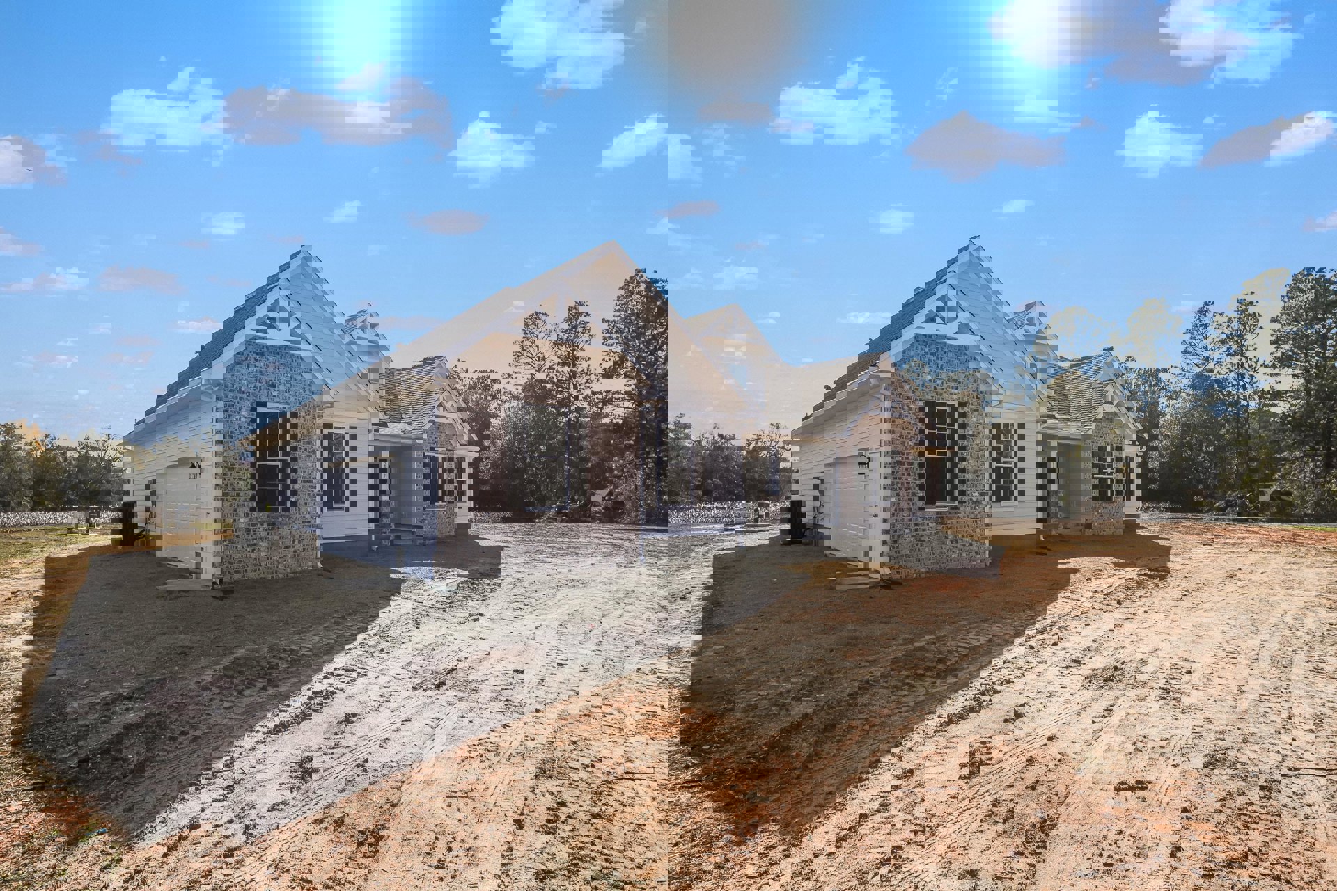 Two-story house with white-framed windows, brick walls, and a black heater beside the exterior; dirt road and trees in front, neighboring homes visible under blue sky with