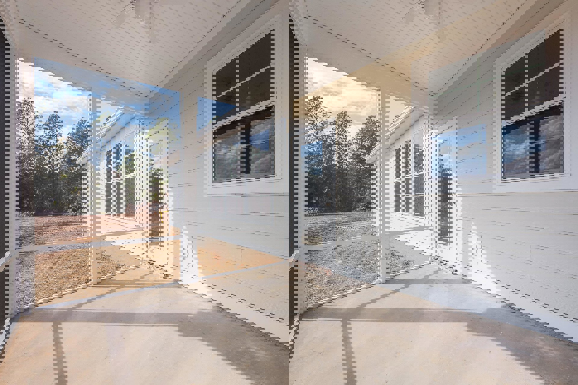 Covered porch with concrete floor, white door featuring glass panel, window reflecting clouds, ceiling fan and light fixture, view of trees through window, light-colored siding