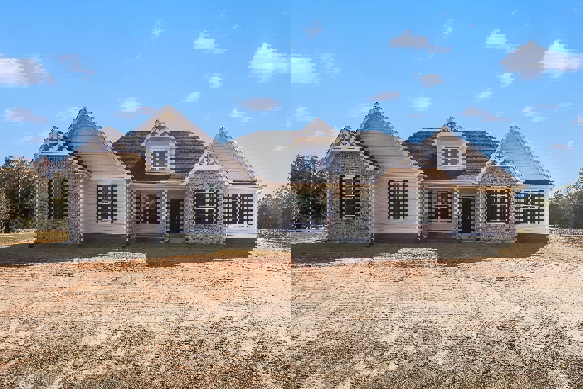 Two-story house with white siding, black front door with glass window, white-framed windows, dormer on gray shingle roof, dirt yard in foreground, sparse landscaping, cloudy sky