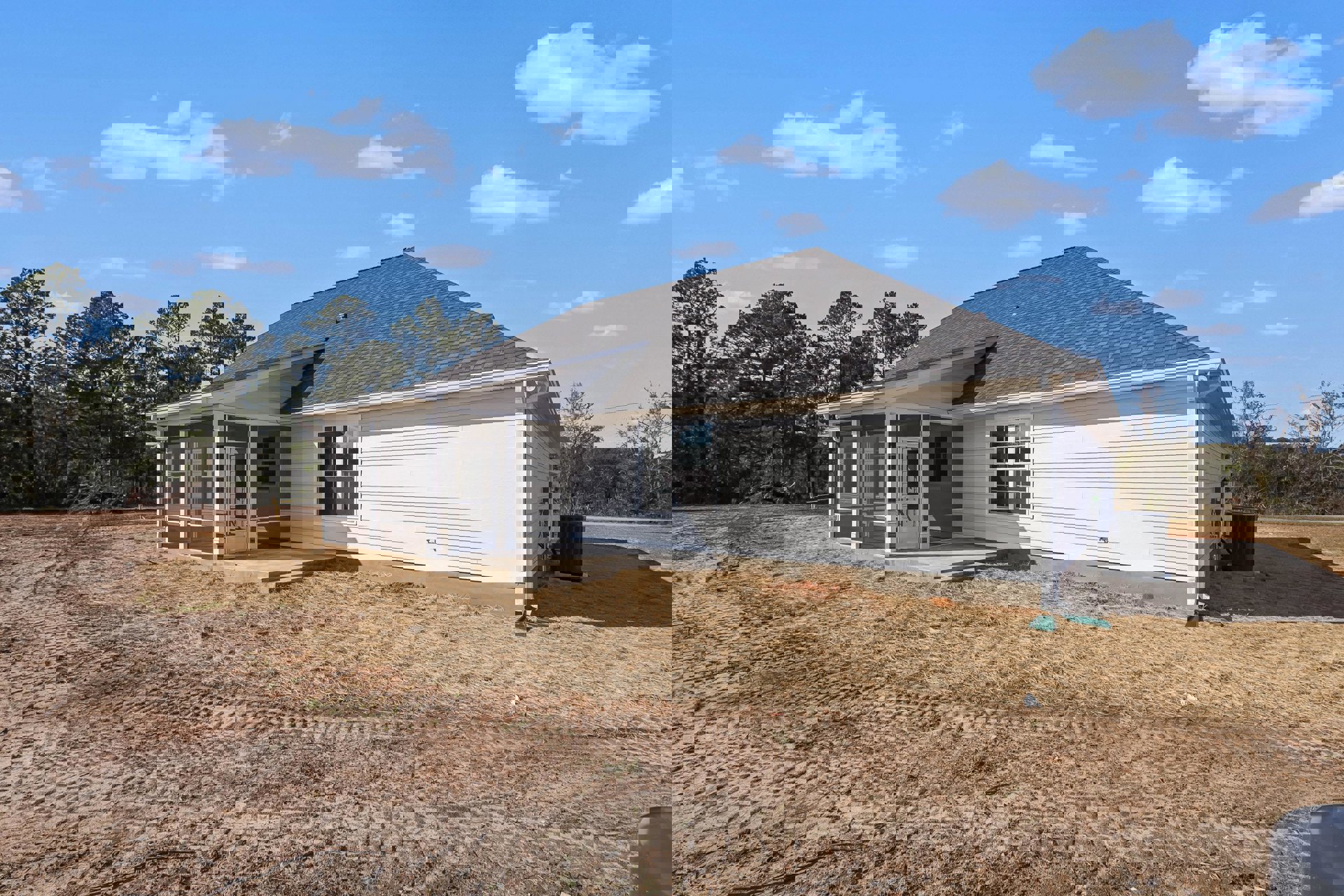 White siding house with covered front porch, unfinished dirt yard, gable roof, and large windows; cloudy sky and sparse trees in background