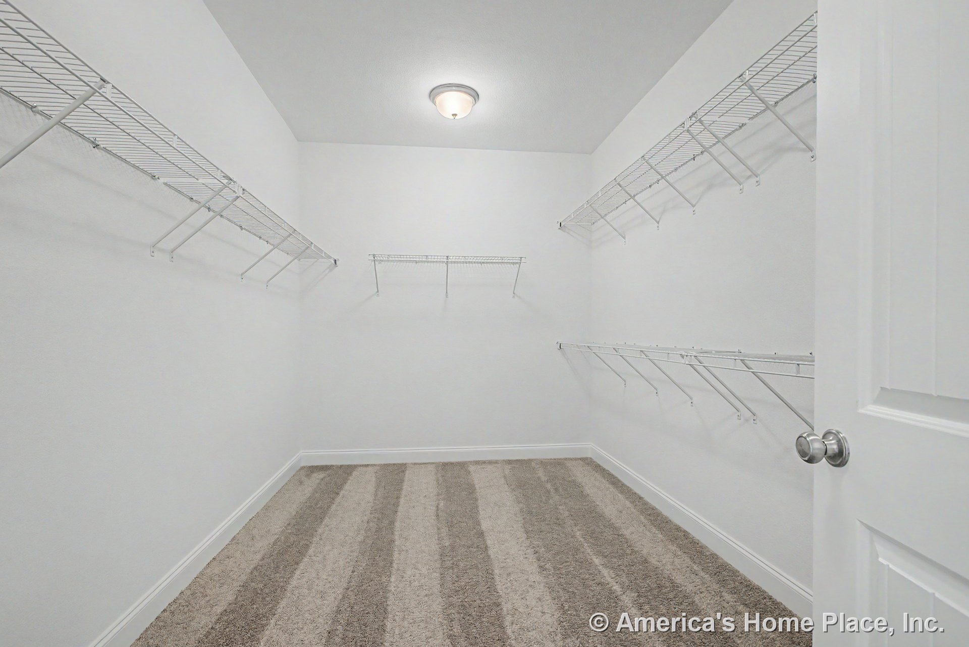 Walk-in closet with multiple white wire shelving units, carpeted floor, flush mount ceiling light, white walls, baseboard trim, and a panel door with round knob.