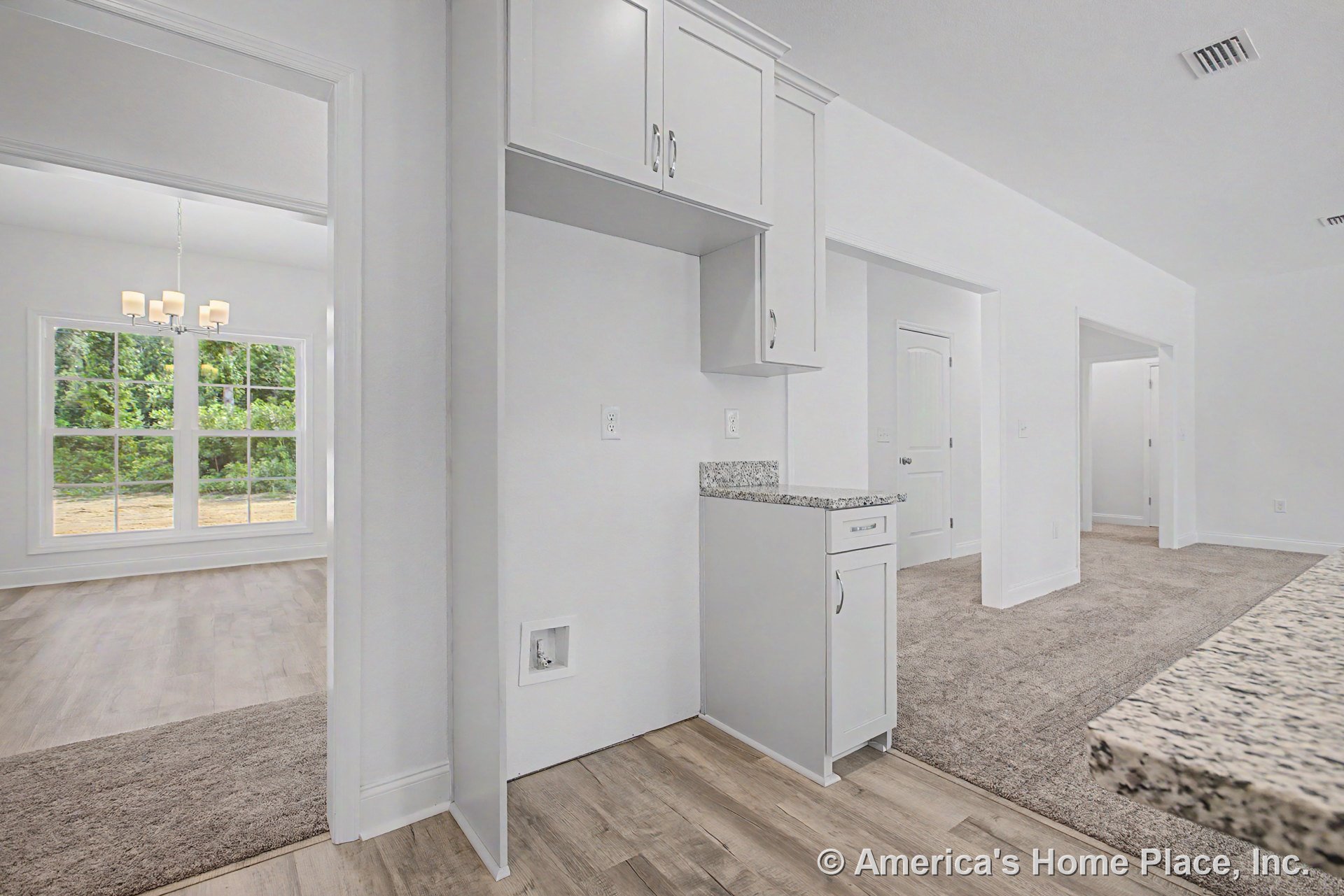 Laundry area with upper built-in cabinetry and granite countertop, adjacent to a carpeted hallway leading into a dining room featuring a chandelier, large double window, wood-look