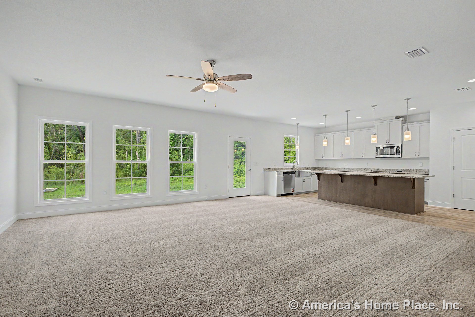 Open concept living area with large triple windows, ceiling fan with light, carpeted flooring, and adjacent kitchen featuring granite island, white upper cabinets, pendant lights