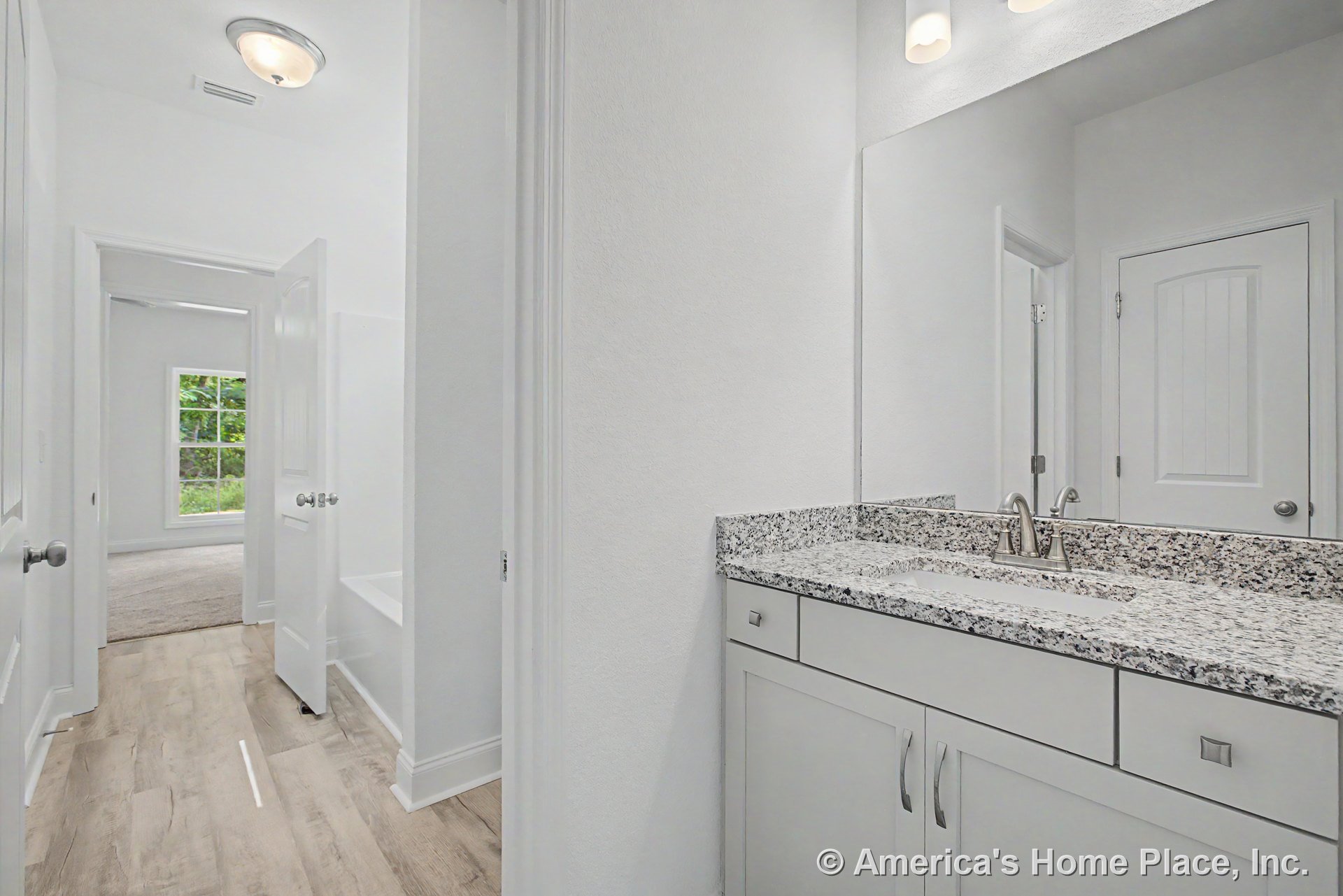 Granite vanity with undermount sink, white shaker cabinets, brushed nickel faucet, large wall mirror, wood-look flooring, recessed panel doors, window, and modern light fixture.