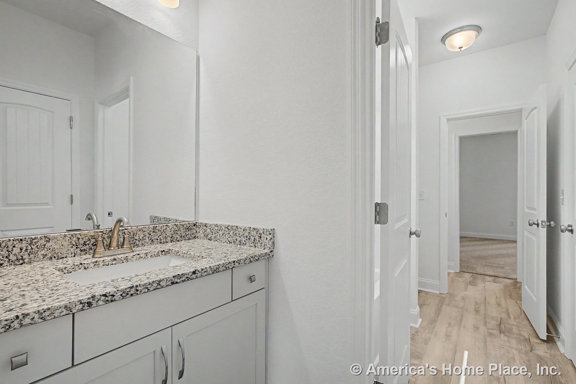 Granite bathroom countertop with undermount sink atop white shaker cabinets, large wall mirror above, hallway with wood-look flooring, panel doors featuring brushed hardware, flush