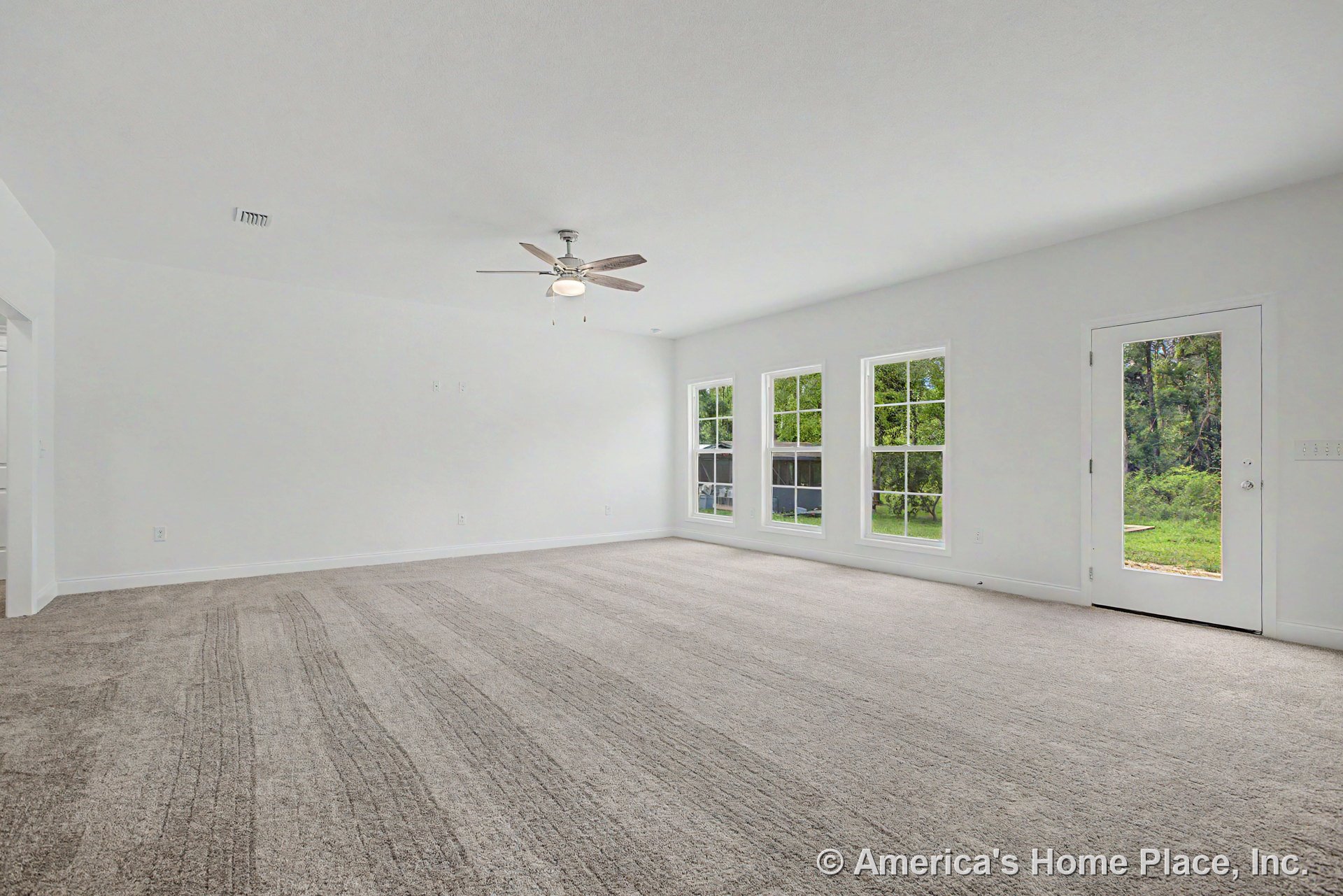 Spacious living room with neutral carpet flooring, three large windows with white trim, ceiling fan and light fixture, smooth white ceiling, and exterior glass door.