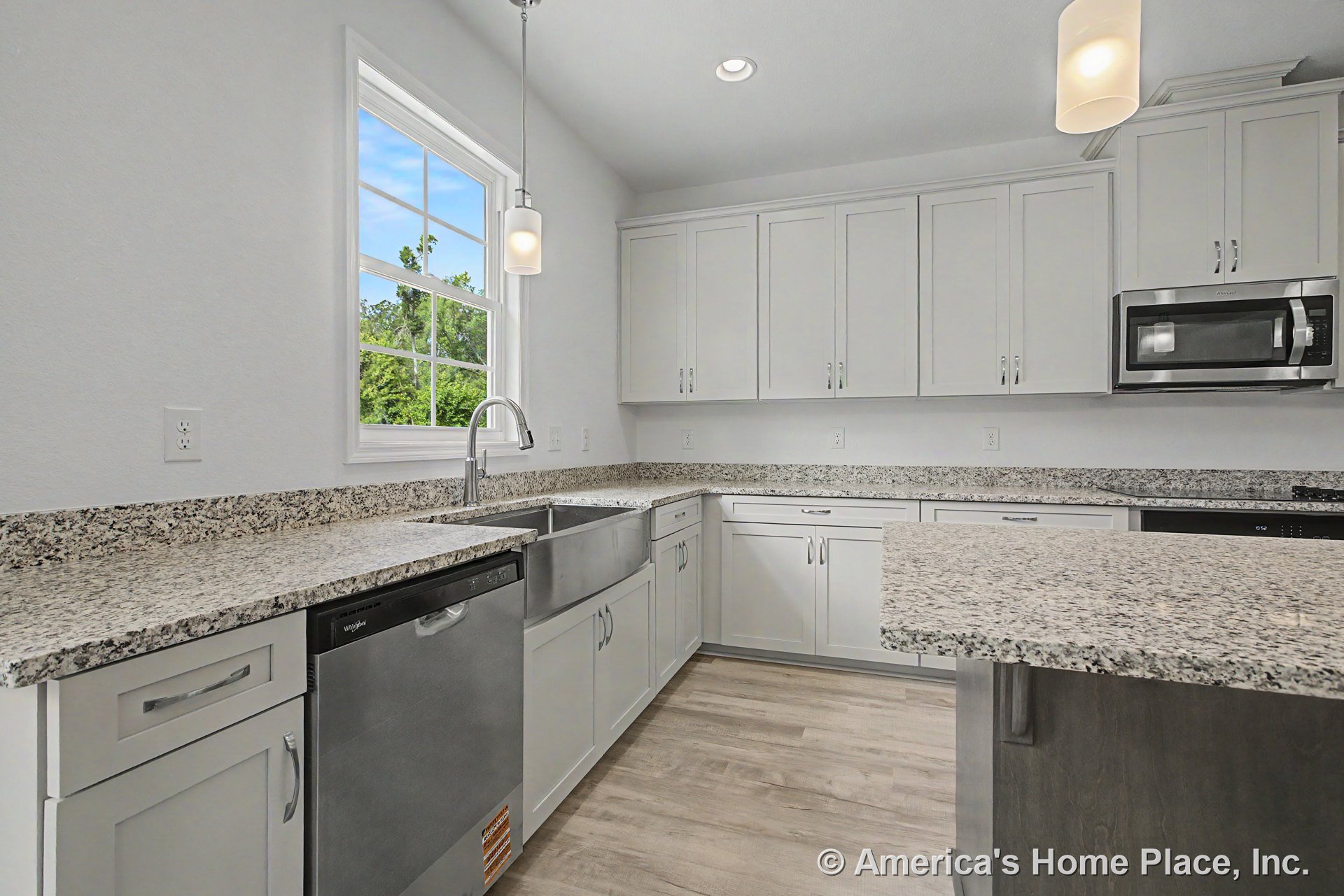 White shaker cabinets with brushed hardware, granite countertops, stainless steel farmhouse sink, built-in microwave, pendant and recessed lighting, large window with decorative