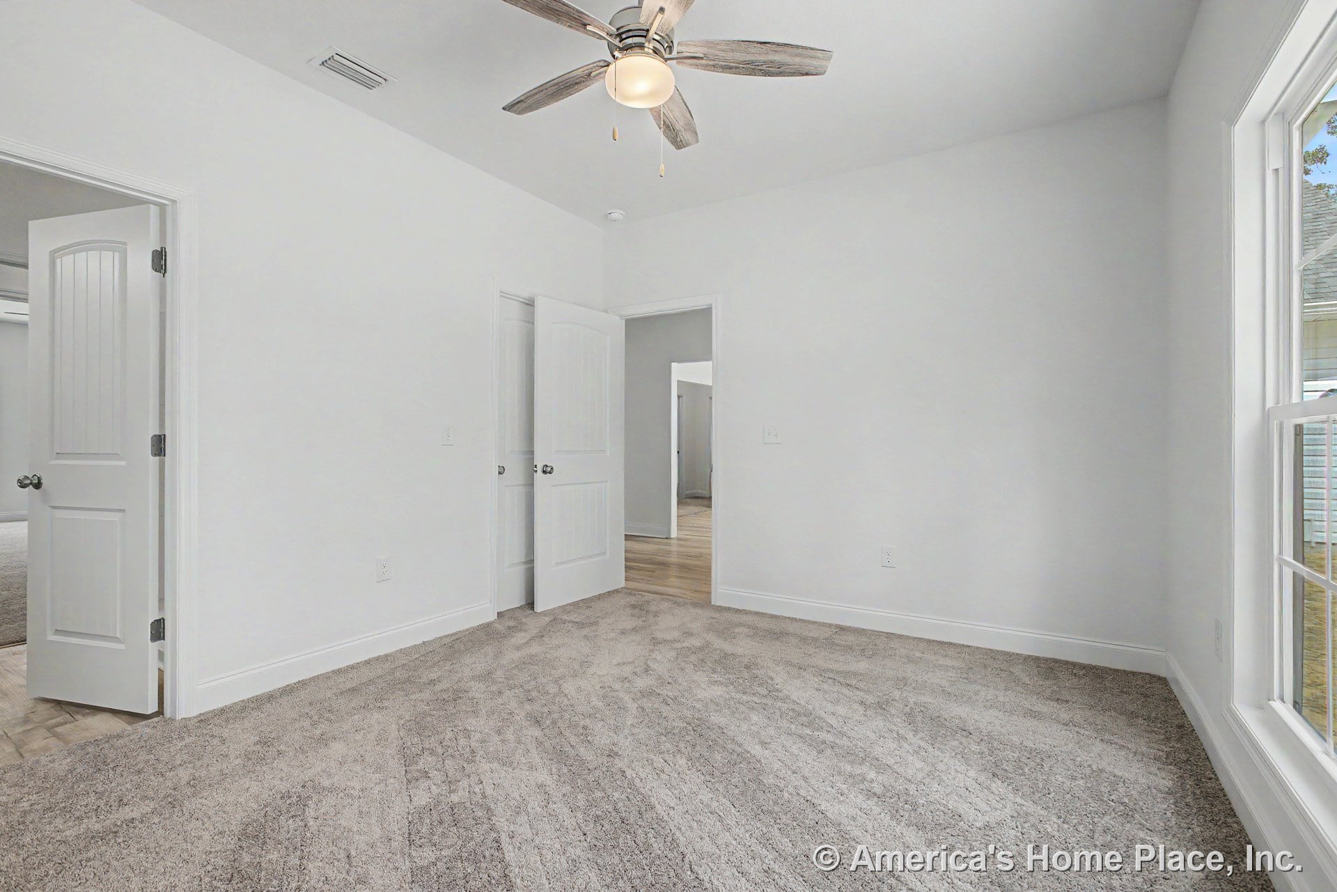 Bedroom with light gray carpet, white walls, large window with white trim, ceiling fan with wood-look blades and integrated light, white paneled doors, baseboard trim, and smooth