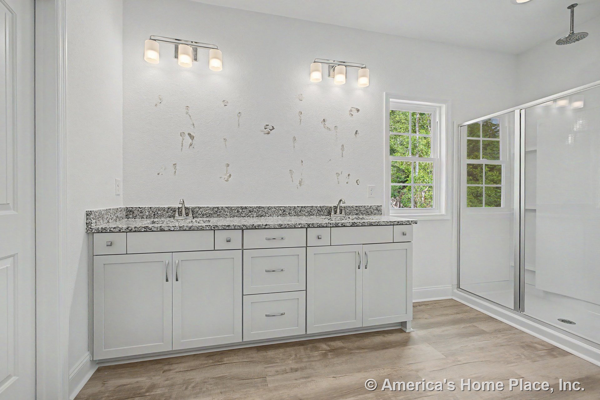 Double-sink vanity with white shaker cabinets and granite countertop, large glass shower enclosure, wood-look flooring, wall-mounted vanity lights, window with trim.