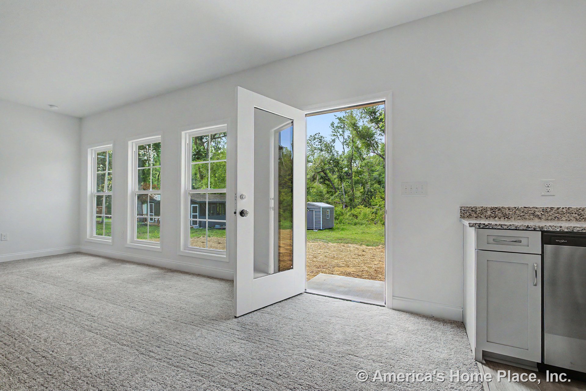 Open living area with three-pane windows, glass exterior door to backyard, granite kitchen countertops, white cabinetry, neutral carpet flooring, and abundant natural light in an