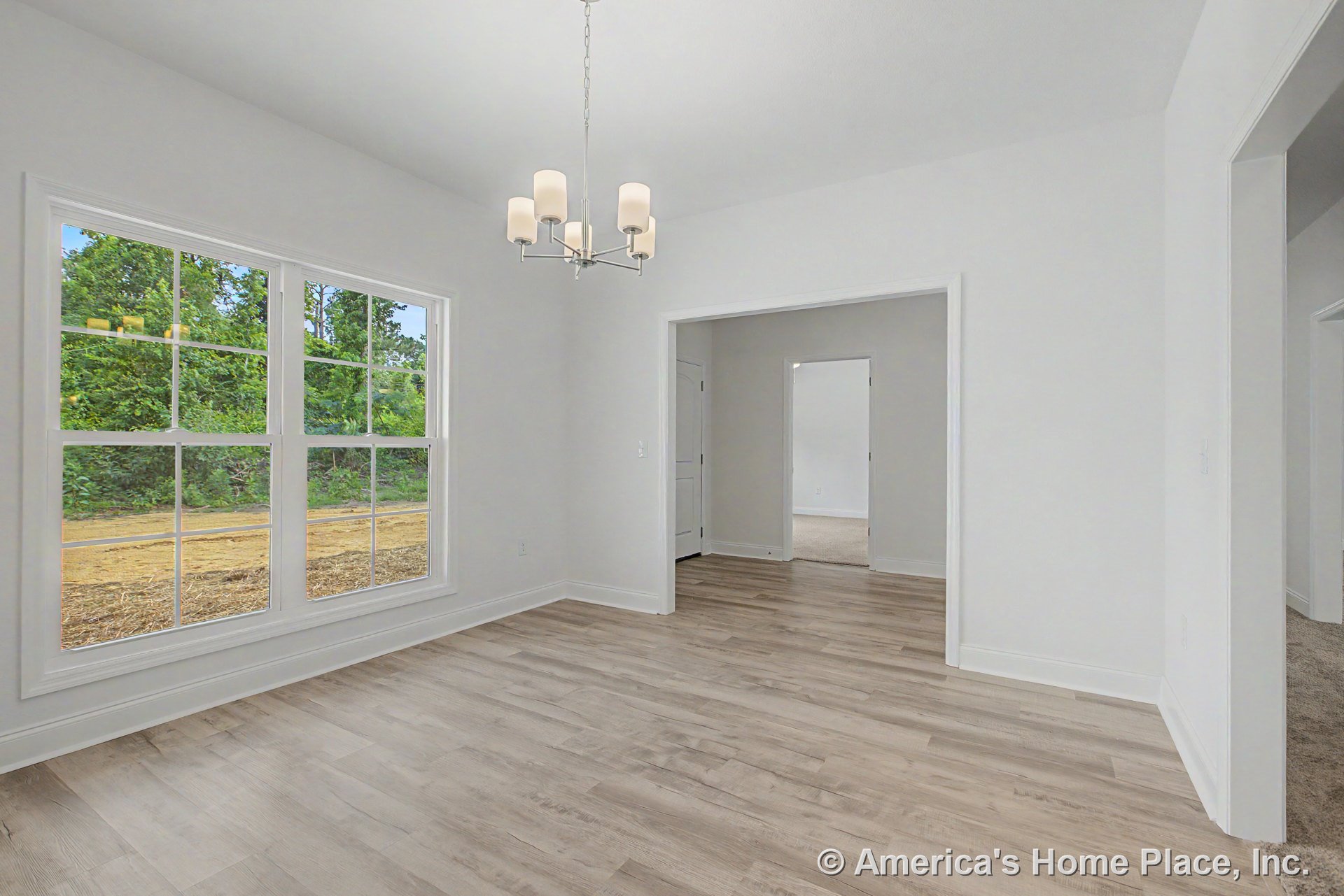 Dining area with large double windows, light wood plank flooring, white trim and baseboards, modern ceiling chandelier, open doorway to adjacent room, and neutral wall color.