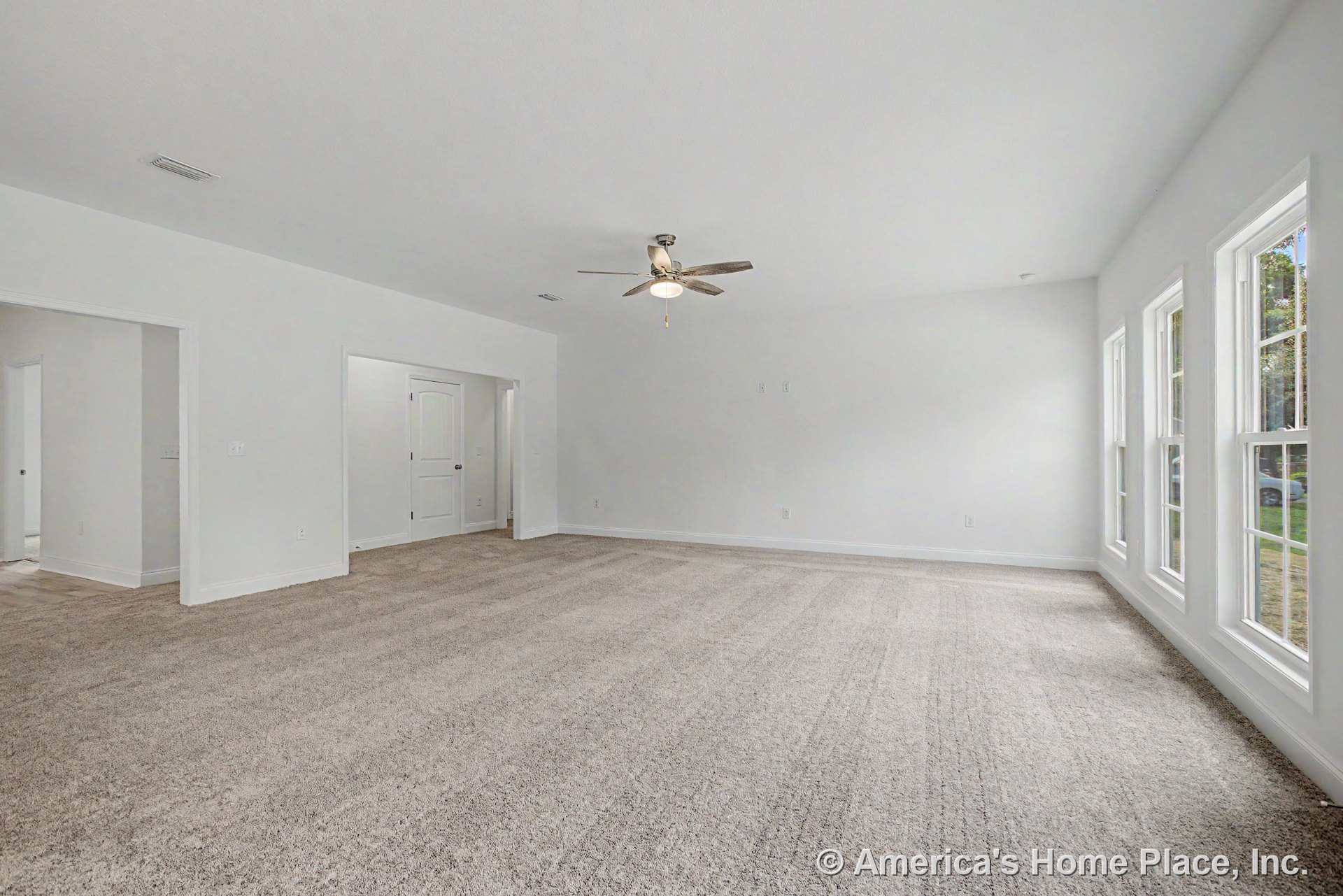 Living room with neutral carpet flooring, three expansive windows allowing natural light, white trim and baseboards, ceiling fan with integrated light fixture, white interior