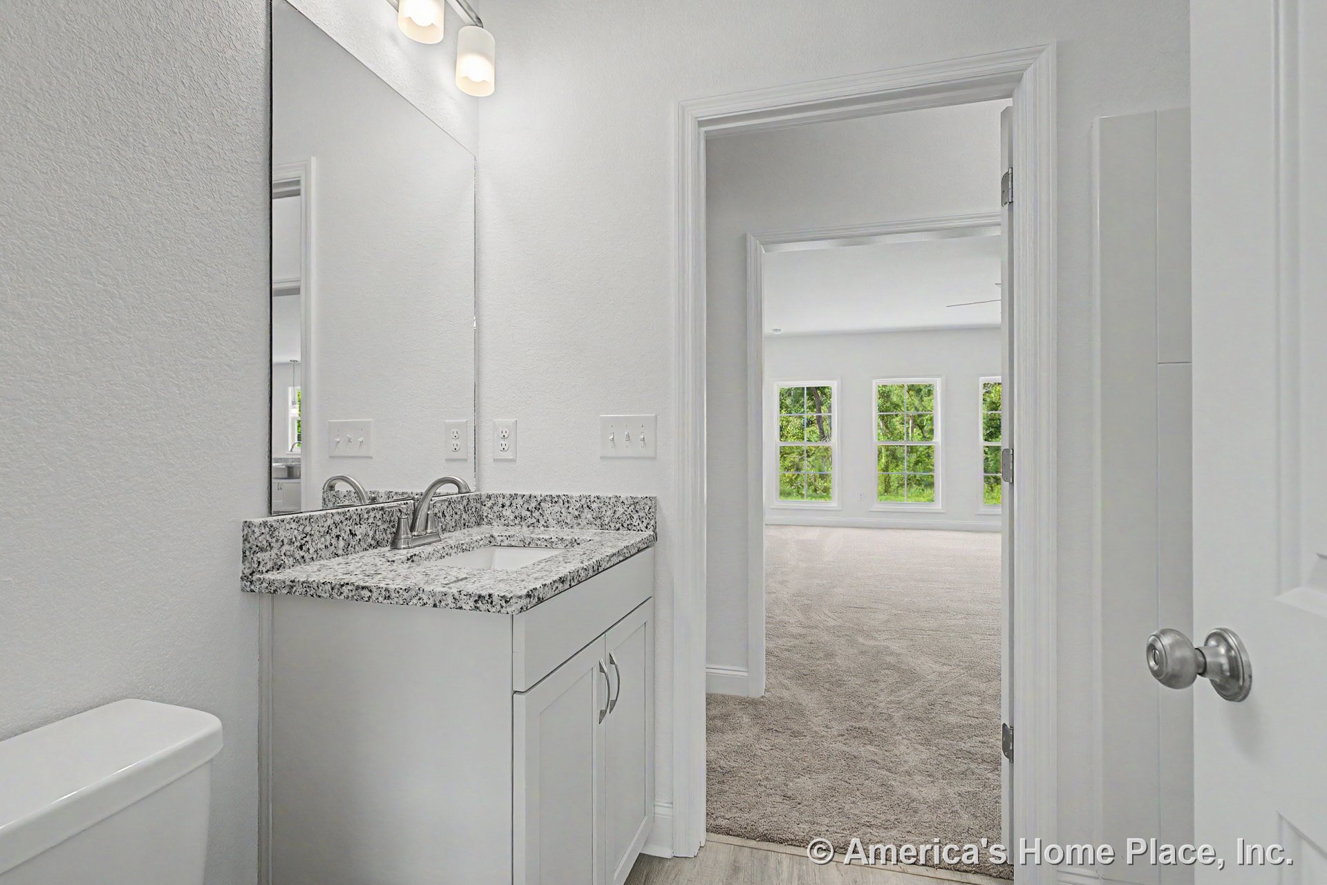 Granite countertop vanity with white cabinetry and large wall mirror, overhead built-in lighting, bathroom opens to carpeted adjacent room featuring multiple large windows with