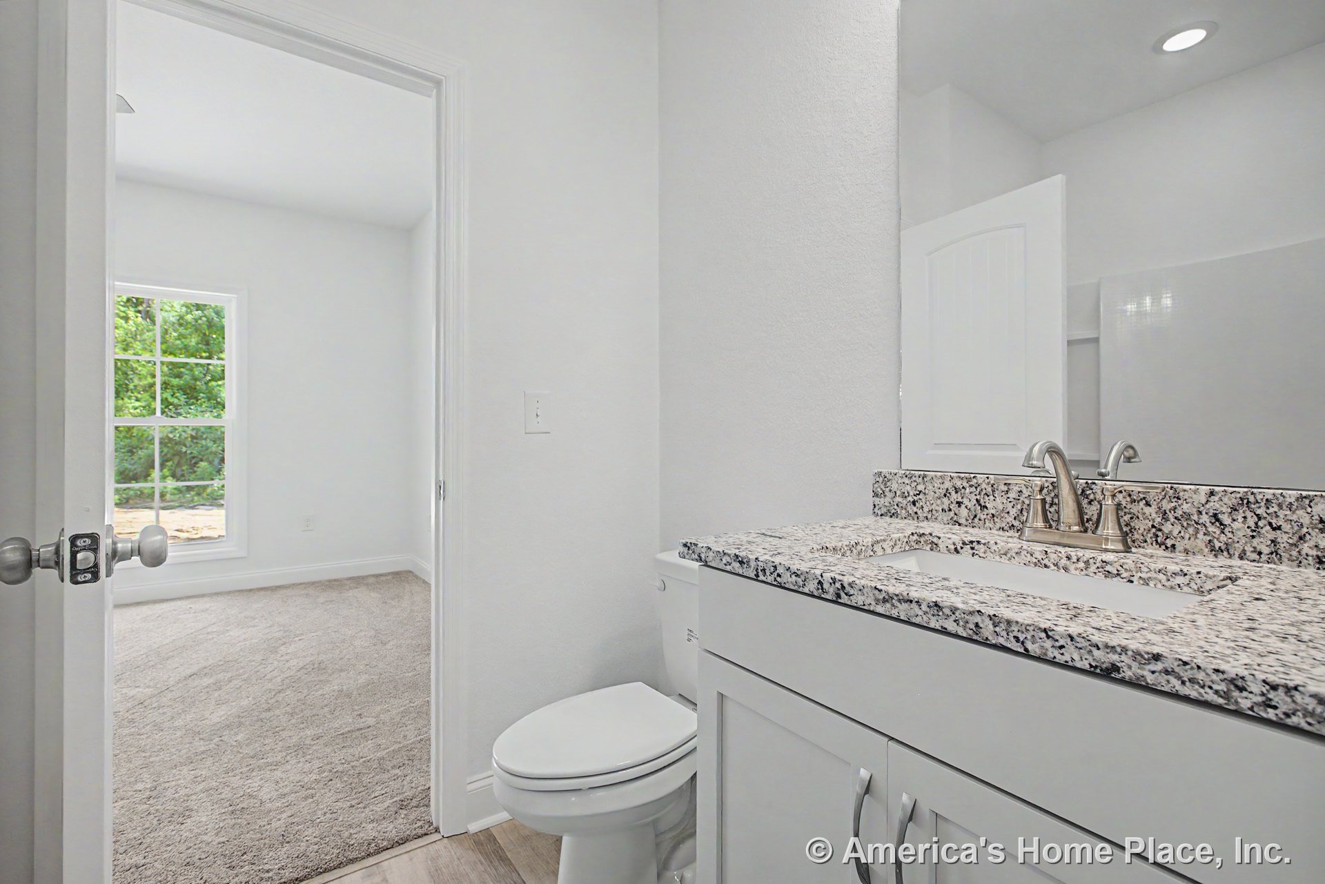 Granite countertop vanity with brushed nickel faucet, white shaker cabinets, large wall mirror, window with grid pattern, recessed lighting, carpeted bedroom visible through open