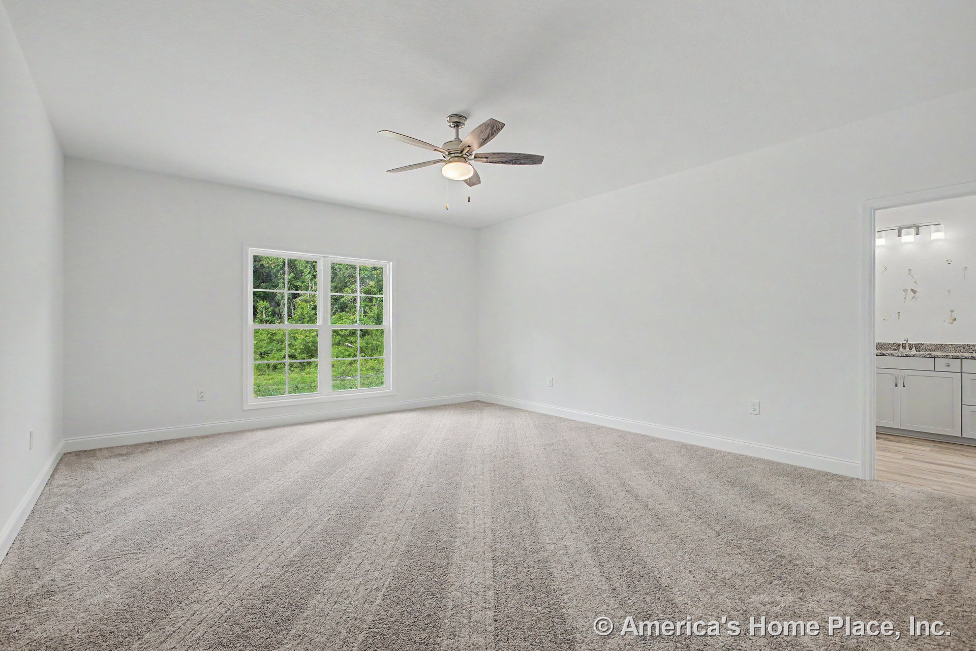 Spacious bedroom with neutral carpet flooring, large grid-pattern window, white painted walls, ceiling fan with integrated light, white baseboard trim, and bathroom entry showing