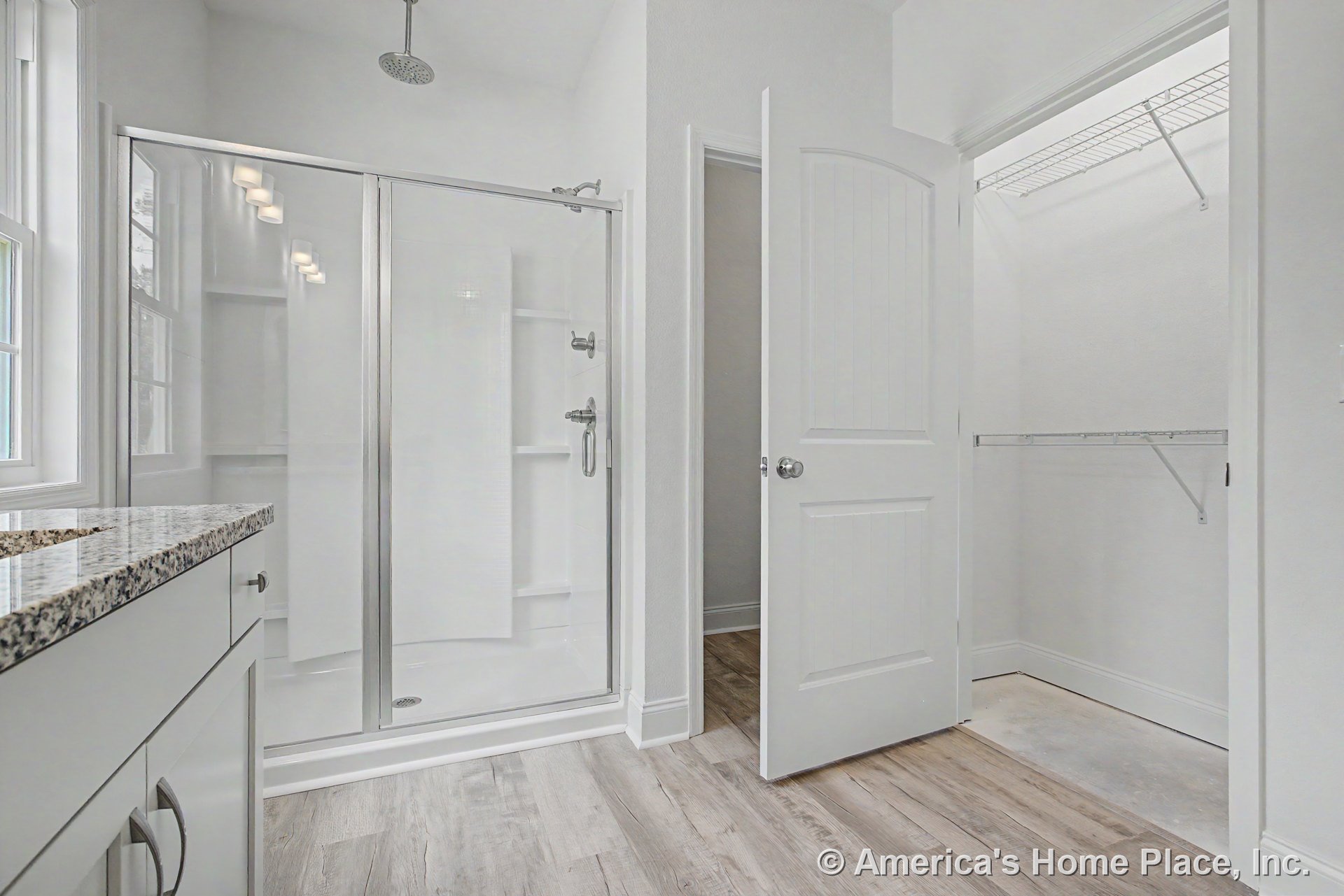 Glass-enclosed shower with overhead fixture, granite countertop vanity with white cabinetry, wood-look flooring, and adjacent walk-in closet featuring wire shelving.