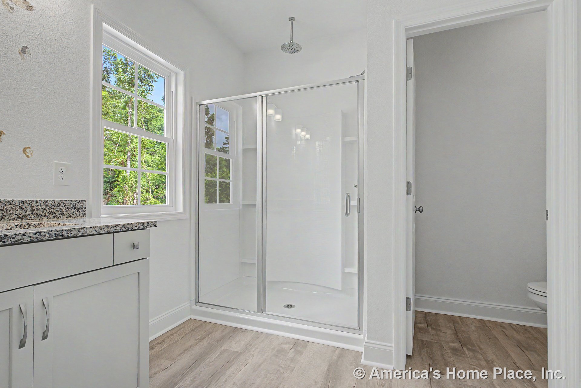 Glass-enclosed shower with ceiling-mounted rain showerhead, granite countertop vanity with white shaker cabinetry, double-hung window, wood-look flooring, white walls, and trim.