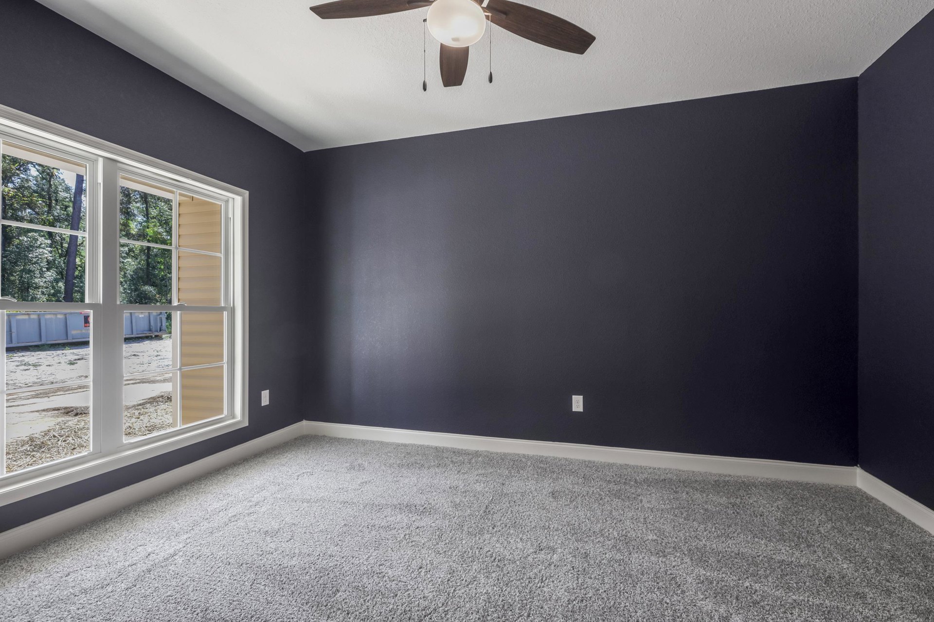Neutral-toned carpeted room with white walls, ceiling fan with light fixture, large window overlooking leafy trees