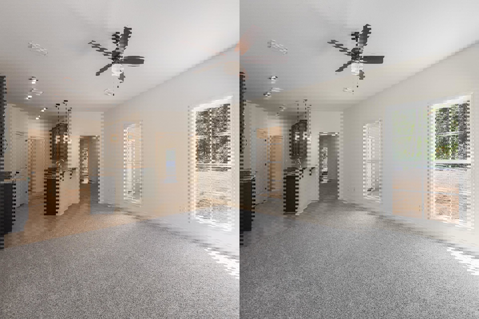 Open-concept living room with beige carpet, ceiling fan with light, white door with black knobs, adjacent kitchen, and large window overlooking forest
