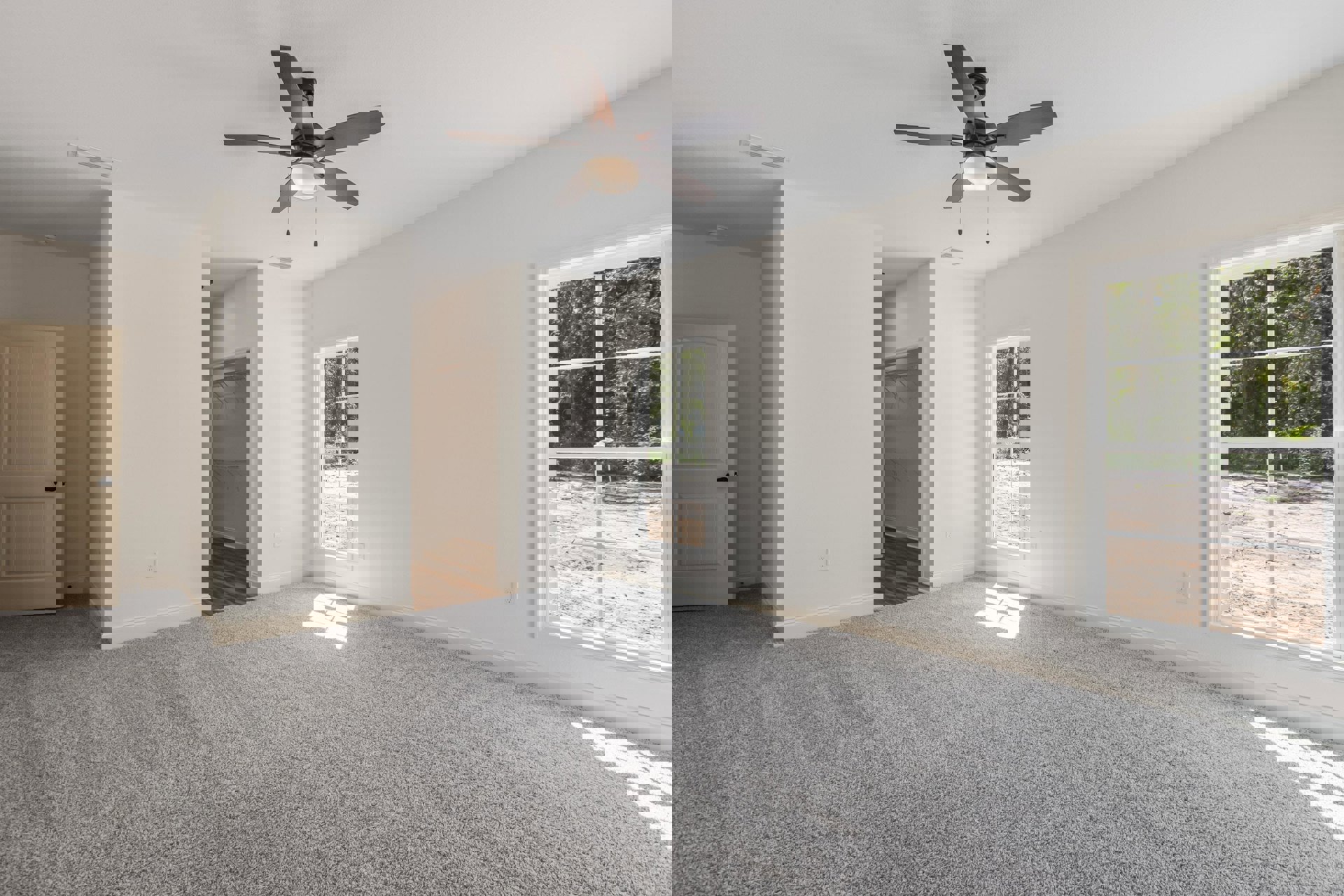 Ceiling fan with light fixture above white carpeted floor, large windows showing trees outside, white door and neutral walls in a bright residential room