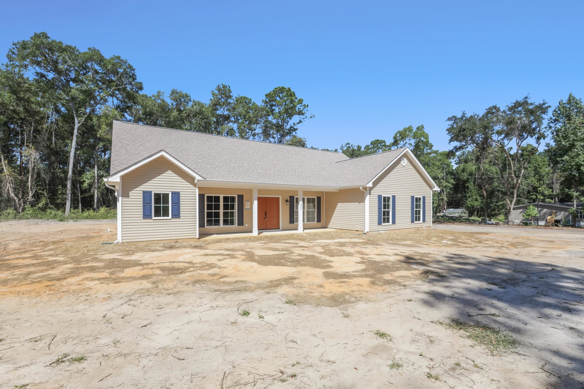 Two-story home with brown front door, white pillars, and white-framed windows, situated on a dirt lot with trees in the background
