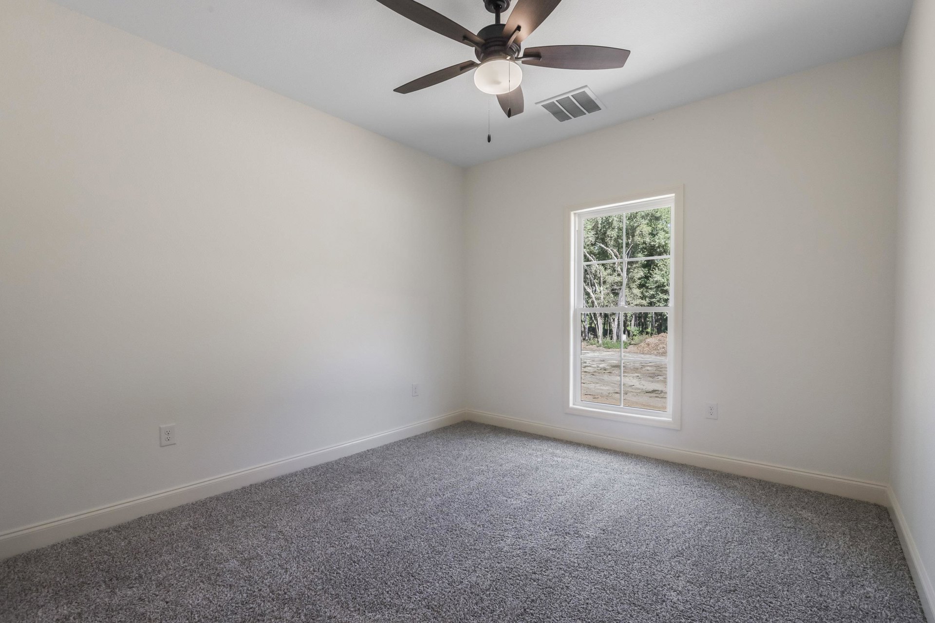 Neutral-toned carpeted room with white walls, ceiling fan and light fixture, rectangular window showing green trees, ceiling vent, and crown molding.