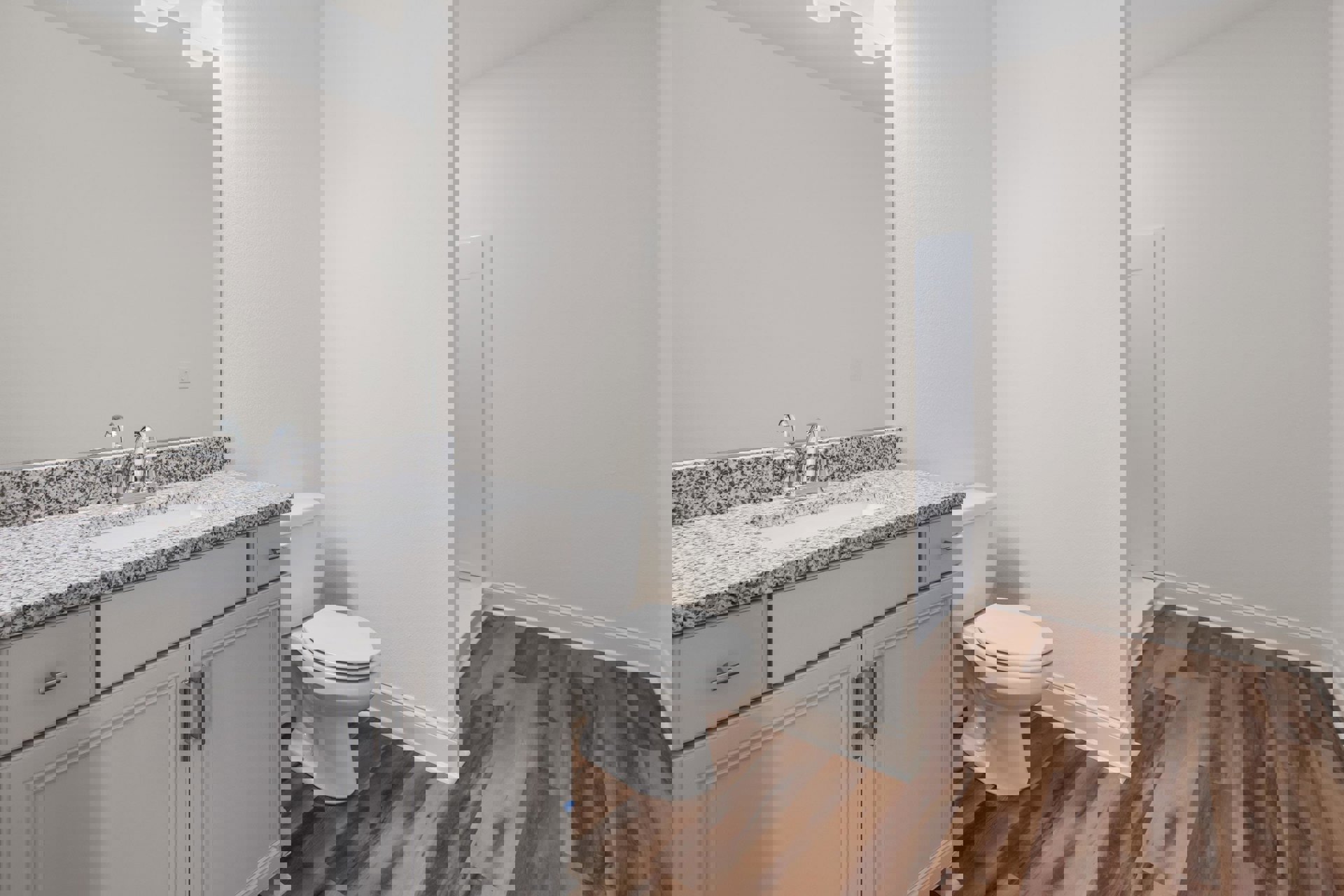 Marble countertop with undermount sink, white toilet with closed lid, wood flooring, white walls, and light-colored cabinetry in a bathroom