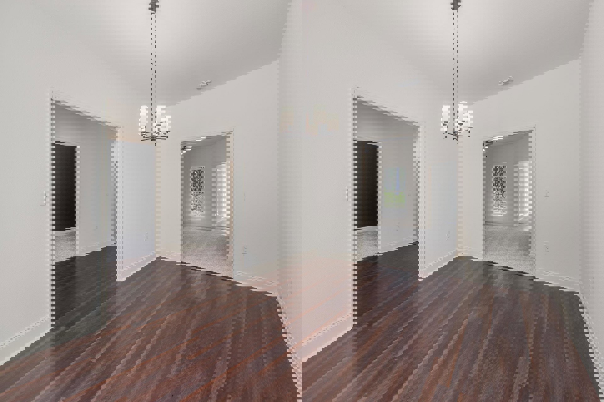 Wood flooring in a spacious room with a modern chandelier, large window revealing trees outside, white plaster walls, and a black rectangular accent object.