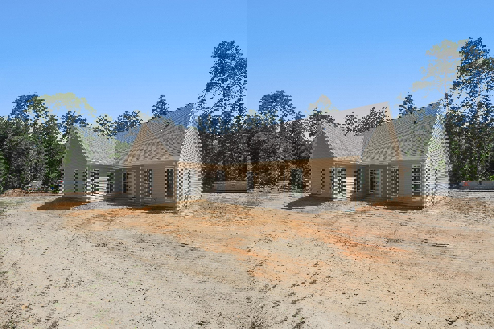 Partially built house with exposed framing, white-trimmed window, front porch, unfinished dirt yard, and mature trees in the background