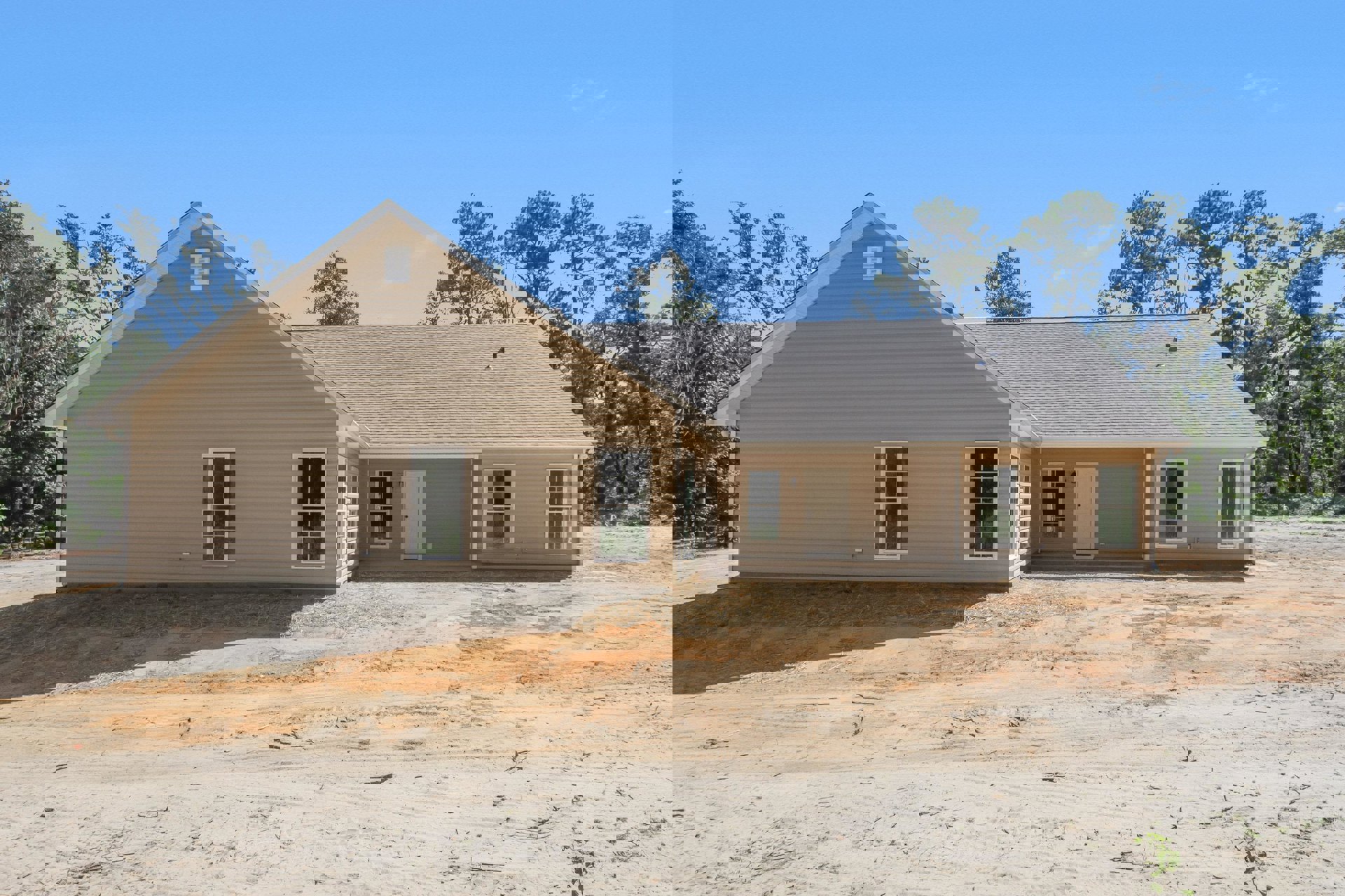 Partially built house with exposed framing, white window and door, black door hardware, dirt yard, gray shingle roof, surrounded by trees under clear sky