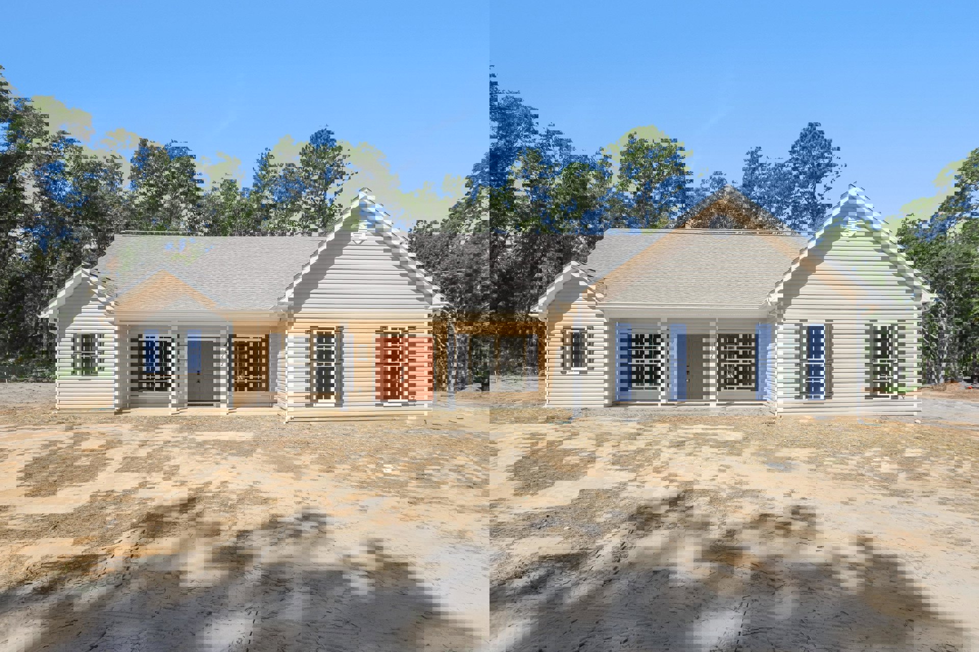Two-story home with white-framed windows, double front door with lock, covered porch, attached garage, dirt driveway, and surrounding trees under a clear sky